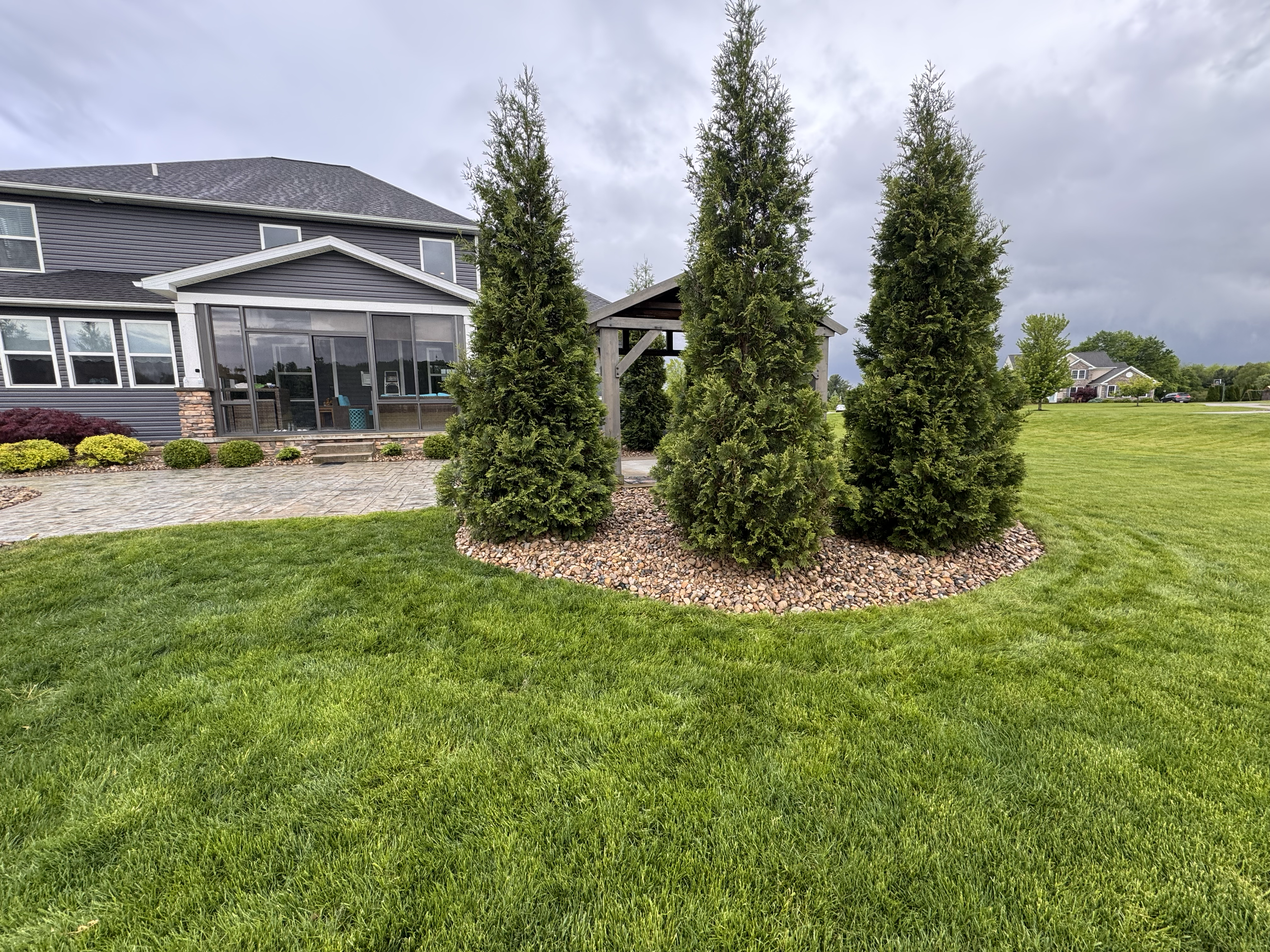 A backyard with three tall evergreen trees surrounded by reddish rocks, a brick pathway, a screened porch attached to a modern house, and a large green lawn with neighboring houses in the background under a cloudy sky.