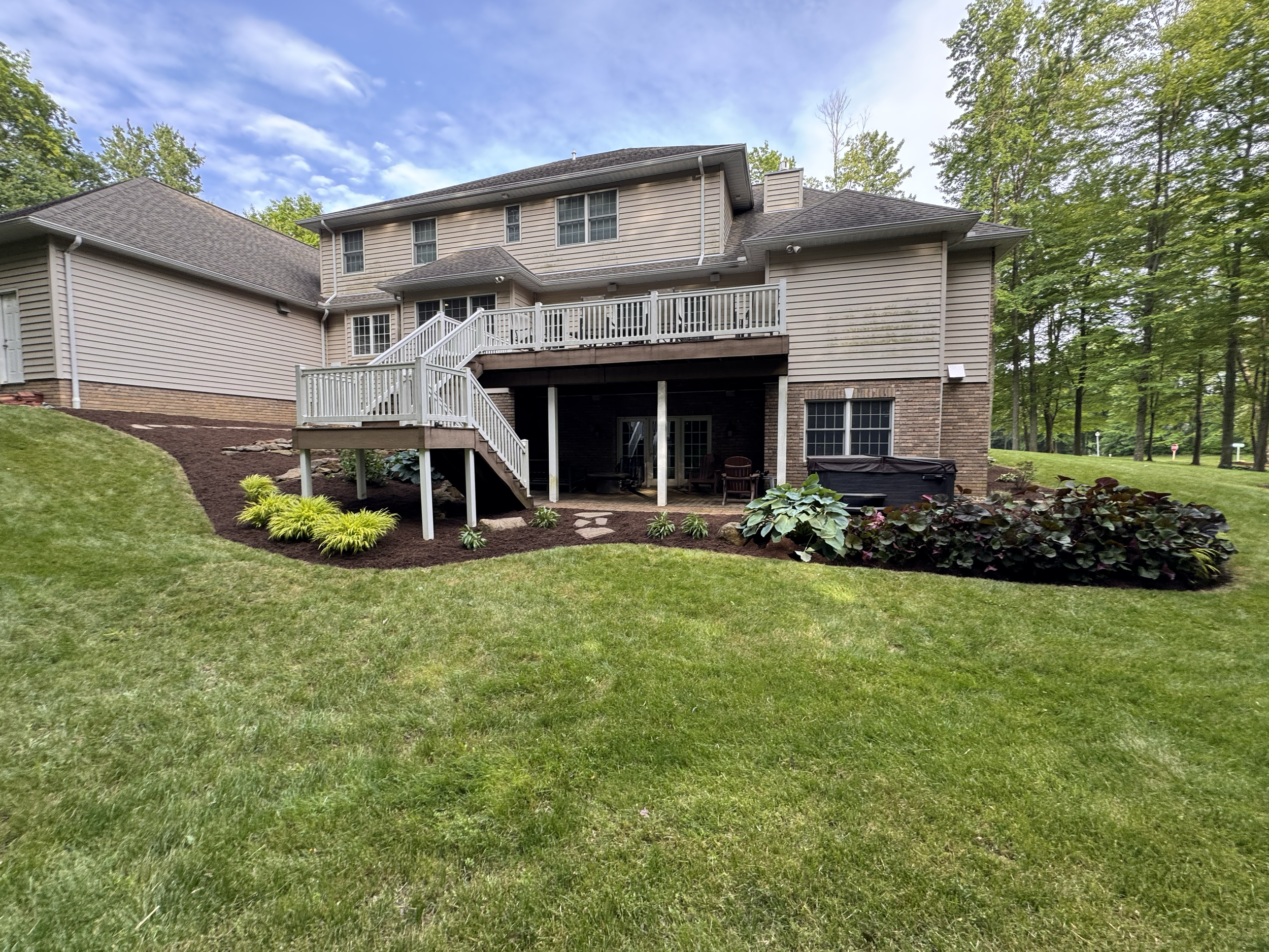 Backyard of a house with a wooden deck, stairs, landscaped garden with plants, and a grassy lawn surrounded by trees under a partly cloudy sky.