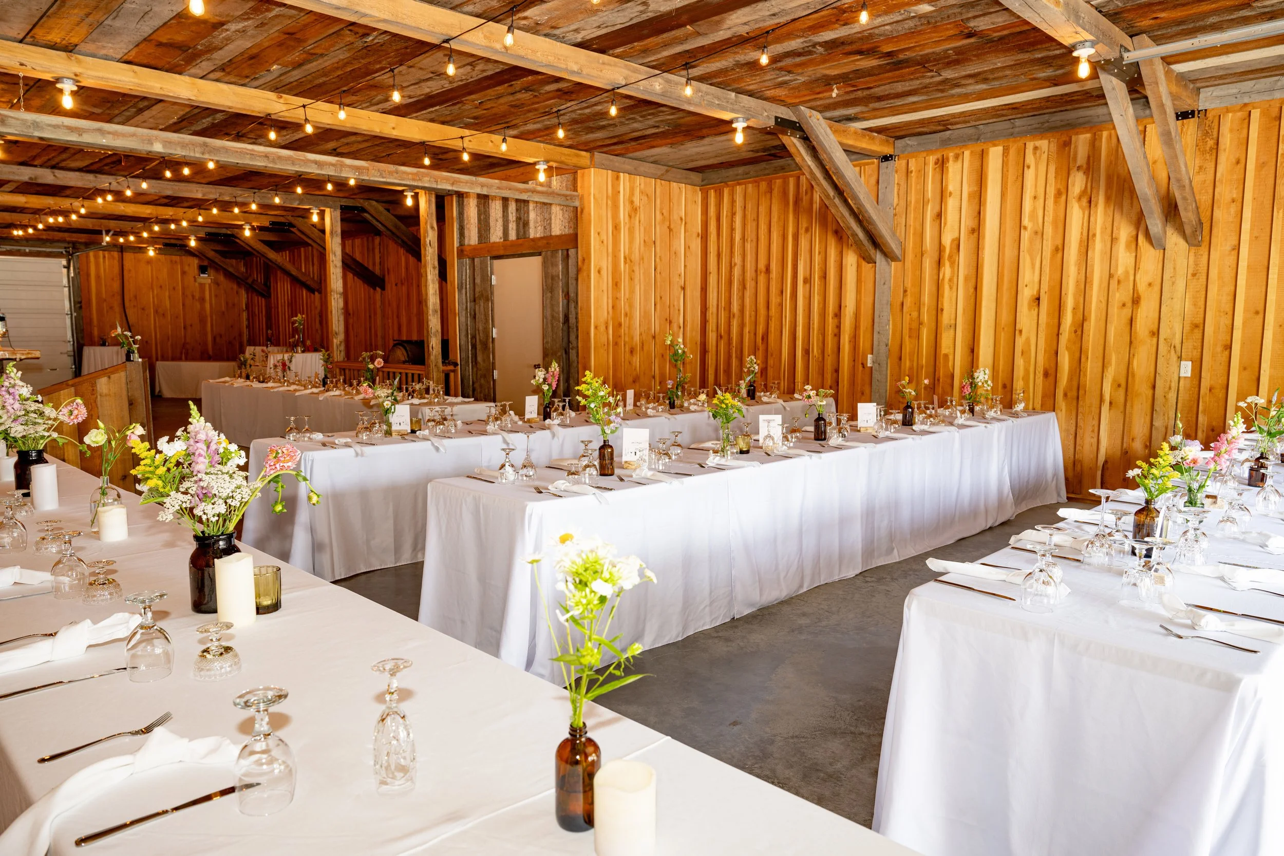 A rustic wedding reception decorated with long tables covered in white tablecloths, floral centerpieces, candles, and neatly arranged tableware in a barn with wooden walls and exposed beams, illuminated by string lights.