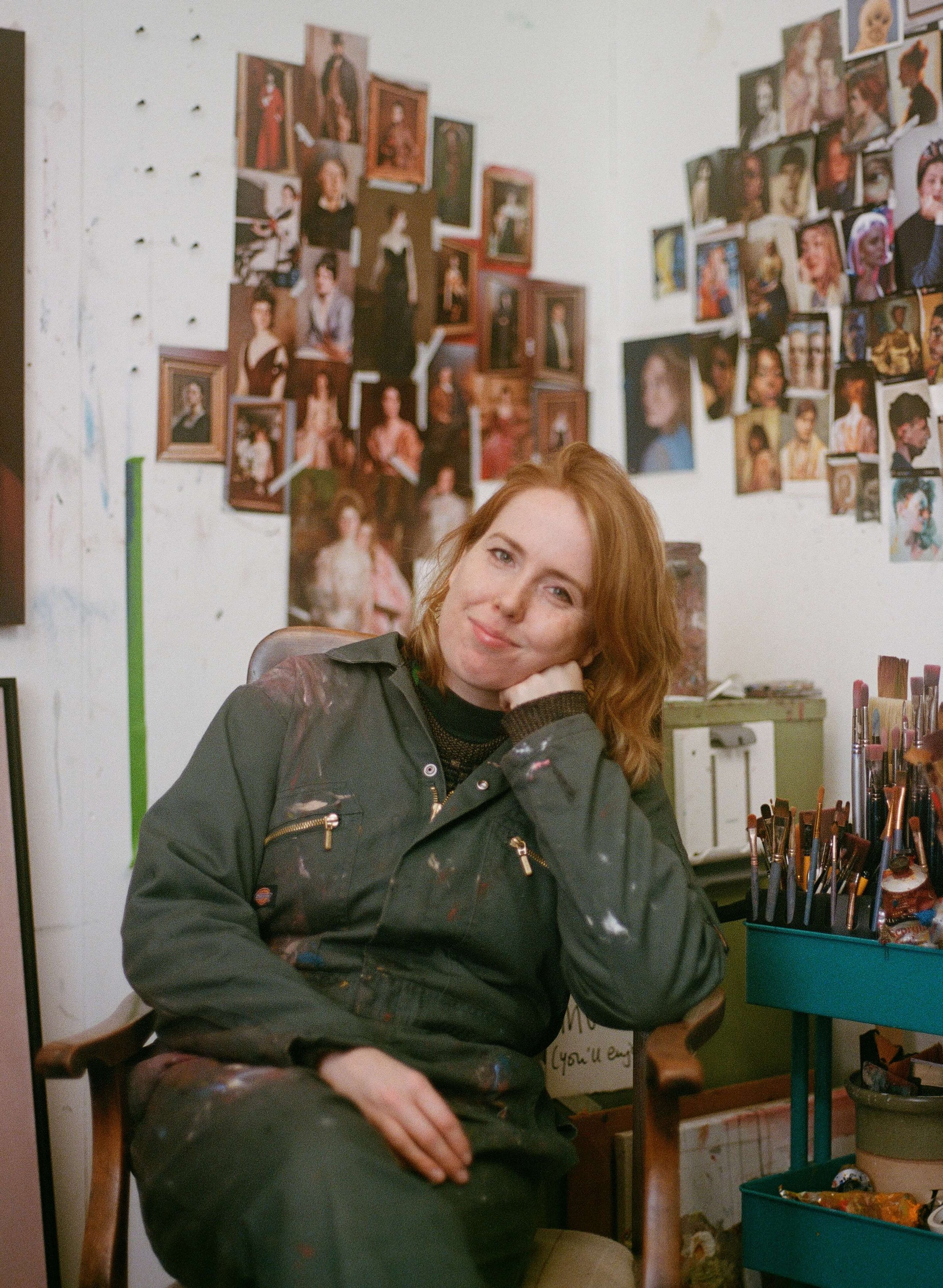 Genevieve sitting in their artist's studio, surrounded by photo collages and paintings of people on the wall.