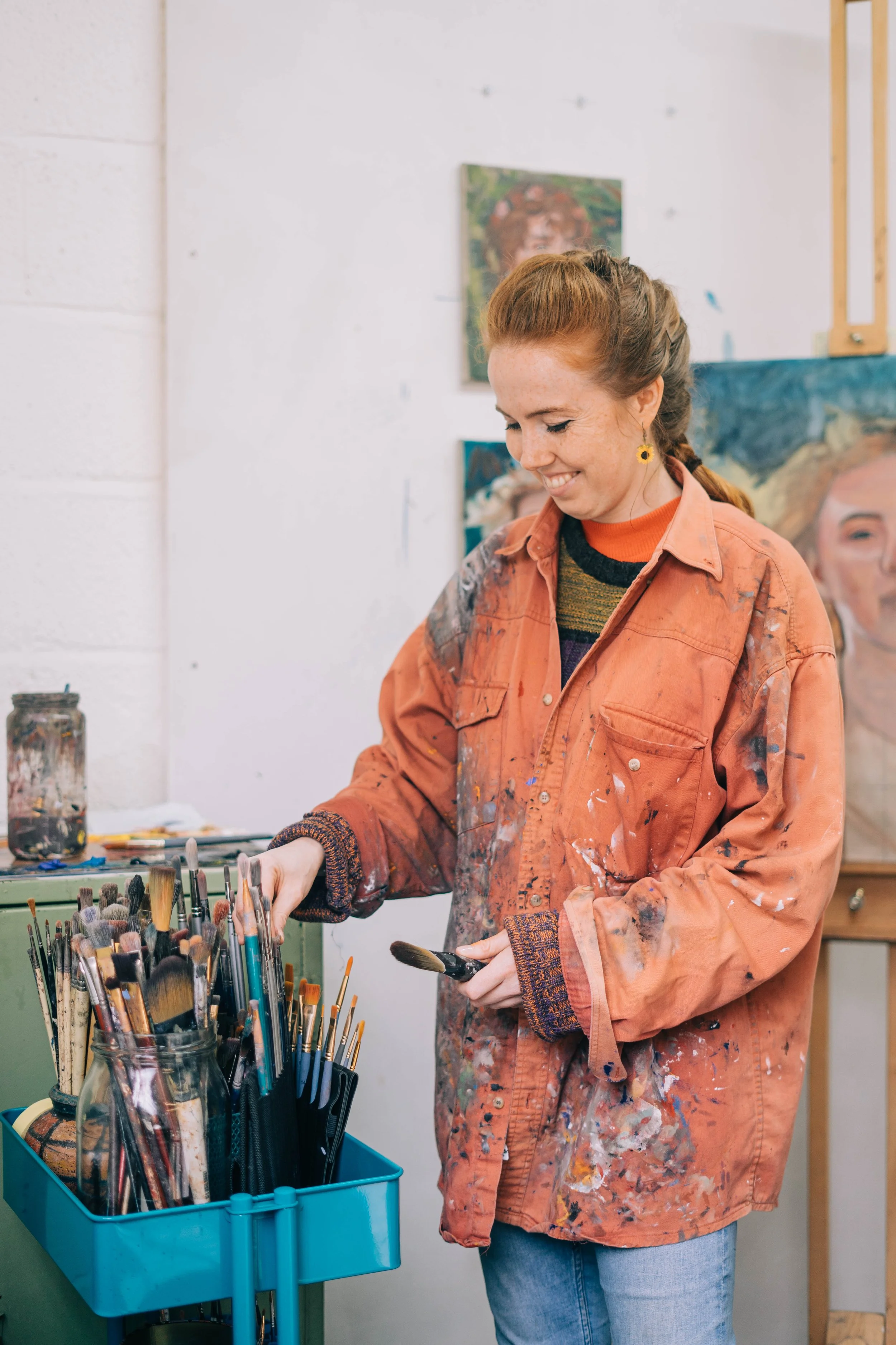 Genevieve Murray Portrait Artist, wearing a paint-stained orange jacket and jeans, smiling while selecting brushes from a container in their art studio in Bristol, South West England.