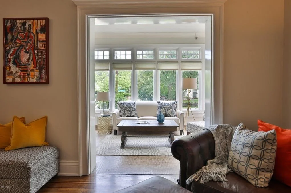 View from a living room into a bright sunroom with large windows, a white sofa, patterned cushions, lamps, and a wooden coffee table.
