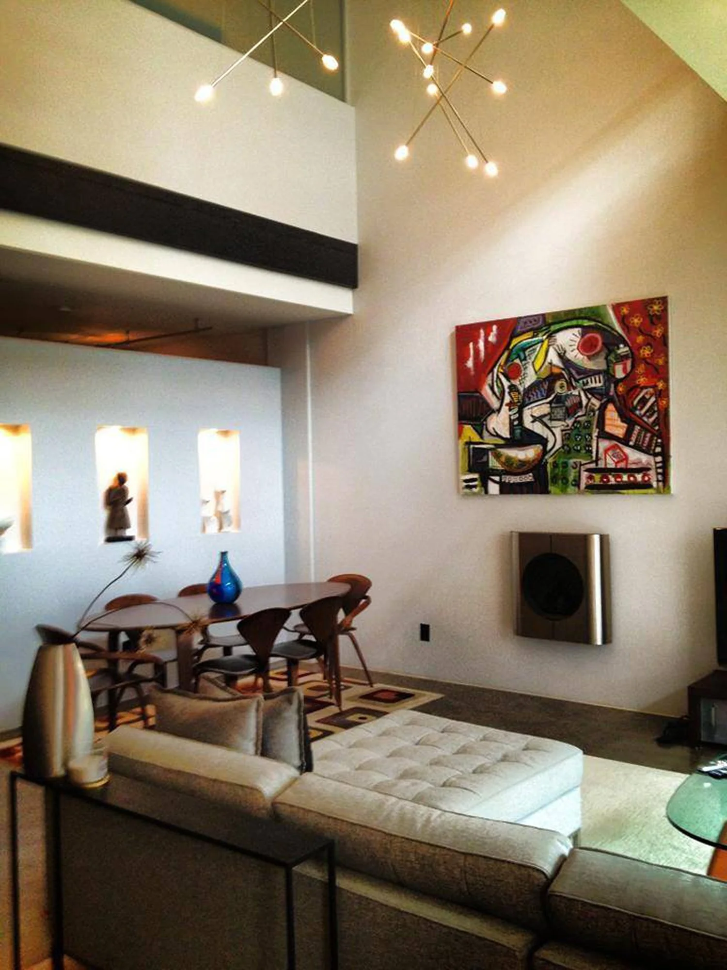 Modern living room with a dining area, abstract colorful painting on the wall, and a contemporary chandelier. Sofa with white and black cushions, glass coffee table, and decorative vases visible.