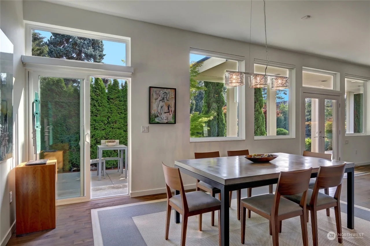Bright dining room with a wooden table and six chairs, large windows showing outdoor greenery, chandelier above the table, and a sliding glass door leading outside.
