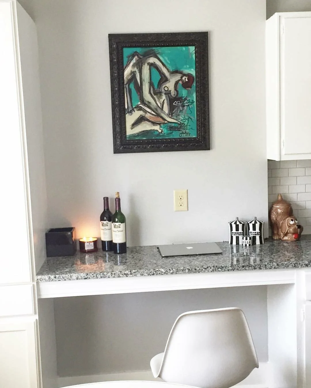 A kitchen counter with a laptop, two wine bottles, a candle, and decorative jars. A framed abstract art piece hangs on the wall above the counter.