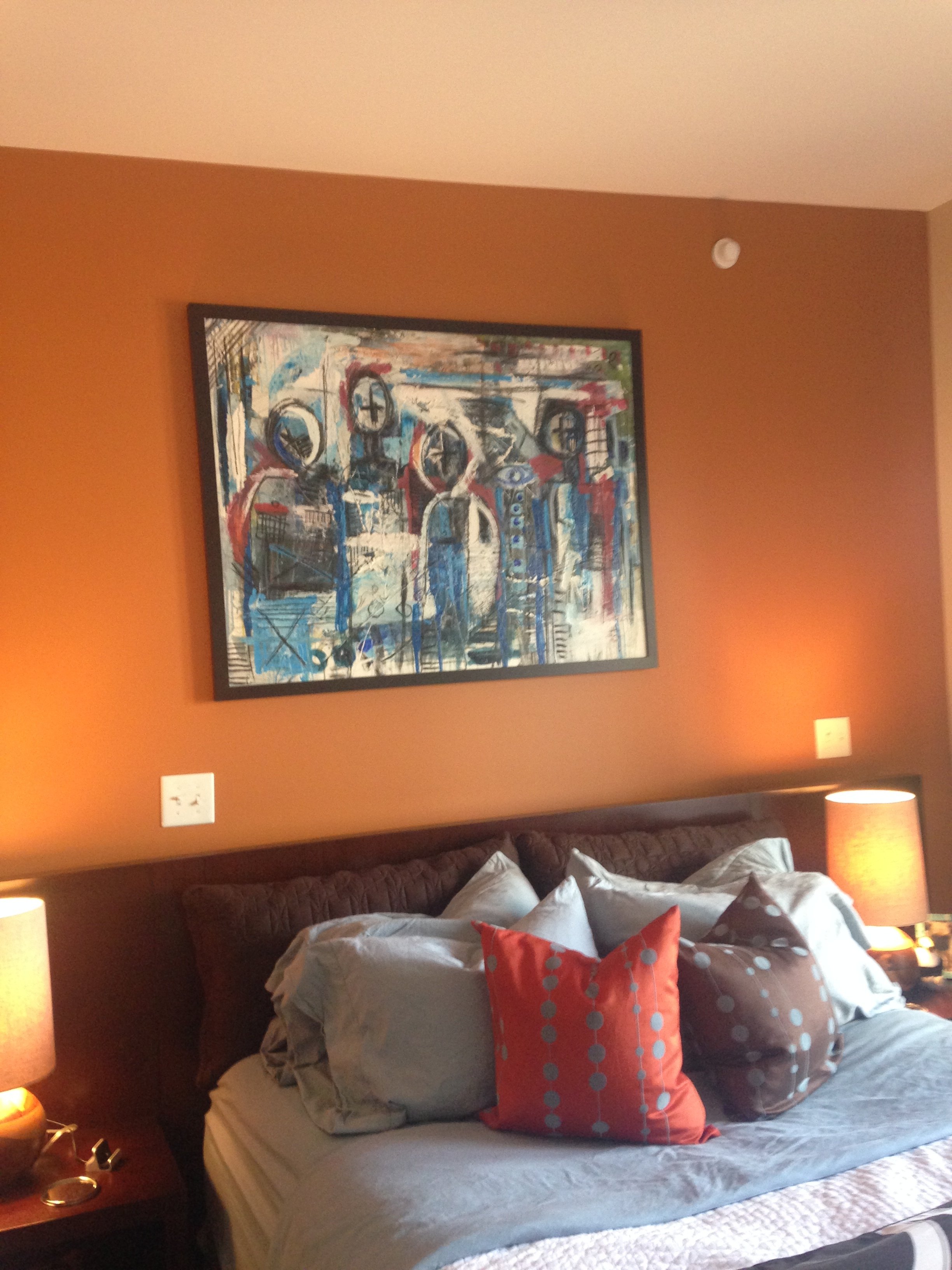 A cozy bedroom with a bed, pillows, and a nightstand, featuring a framed abstract painting on a warm-colored wall and two lamps providing warm lighting.