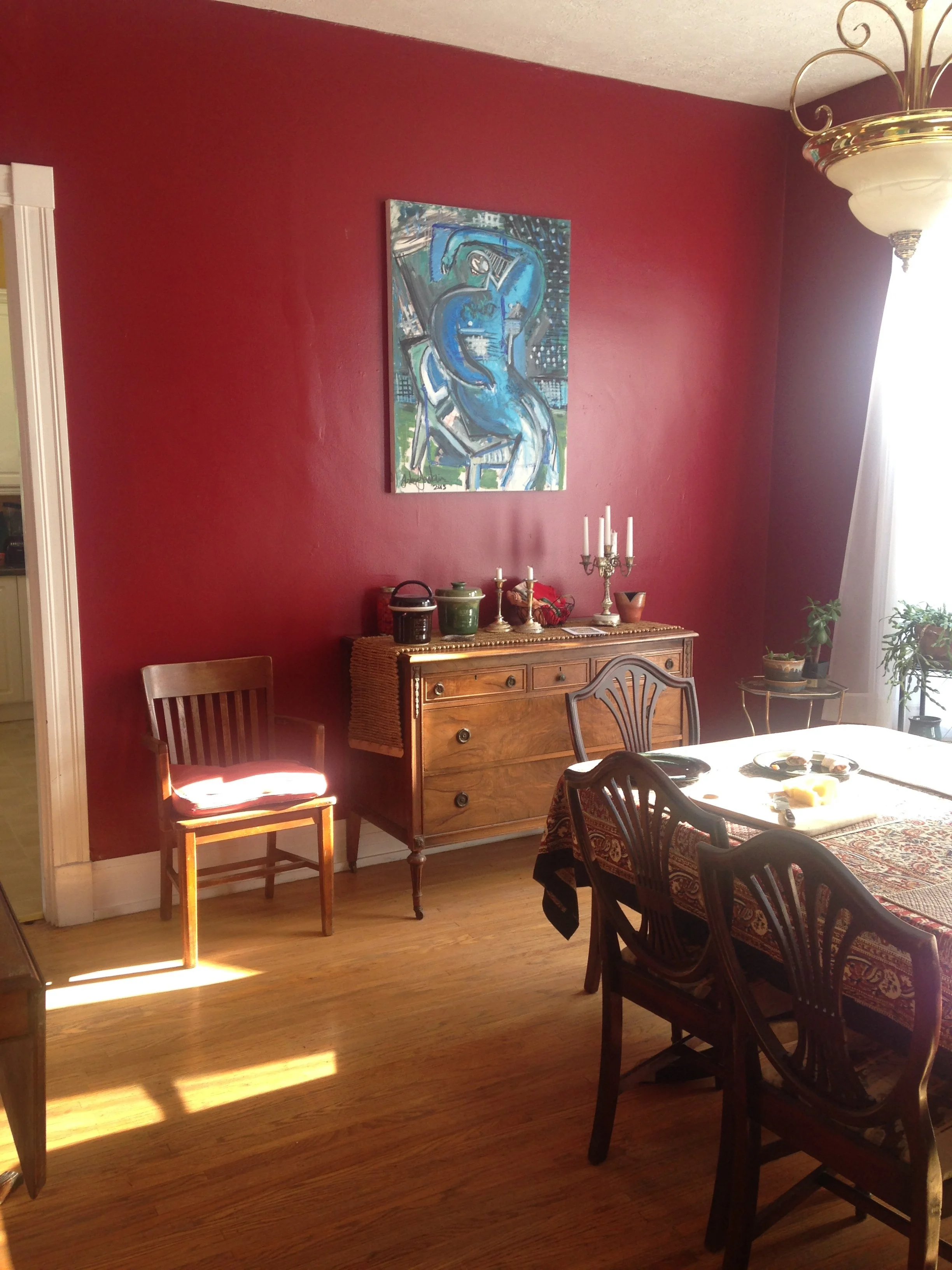 A dining room with a red wall, wooden floor, a wooden sideboard with decorative items, a painting on the wall, and a table with chairs, some with a tablecloth and some without. Light coming through a window on the right.