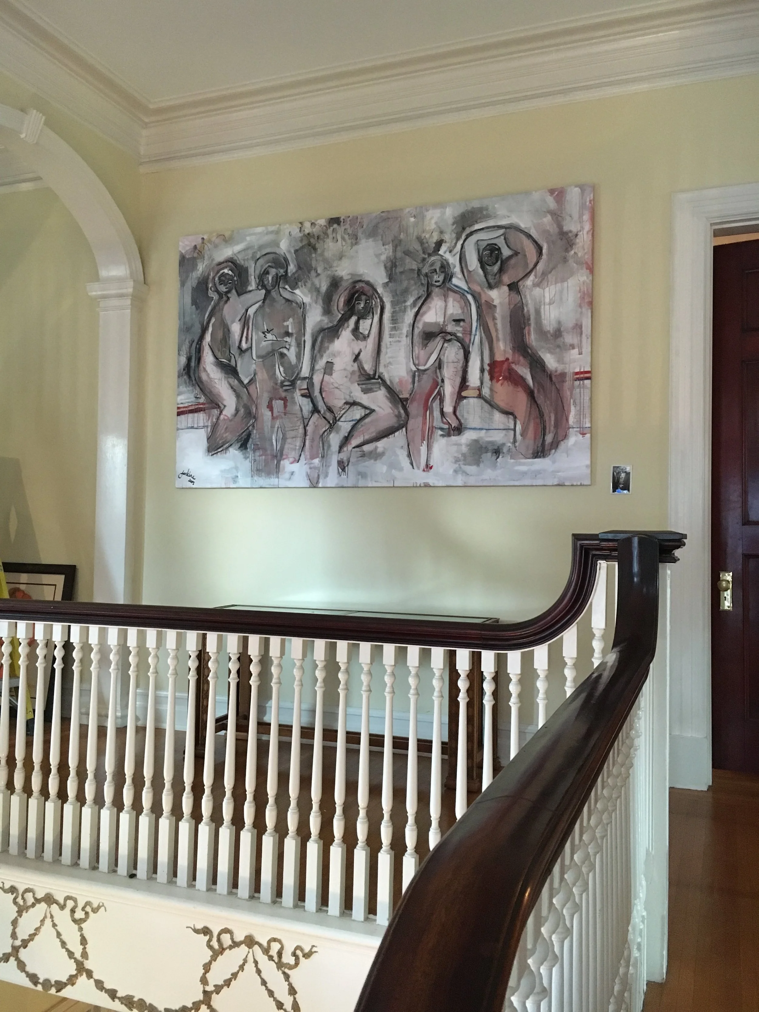 Large modern abstract painting of five women with dark skin tones standing and sitting, painted mostly in shades of gray, black, white, with some pink and red accents, hanging on a yellow wall in a house interior.