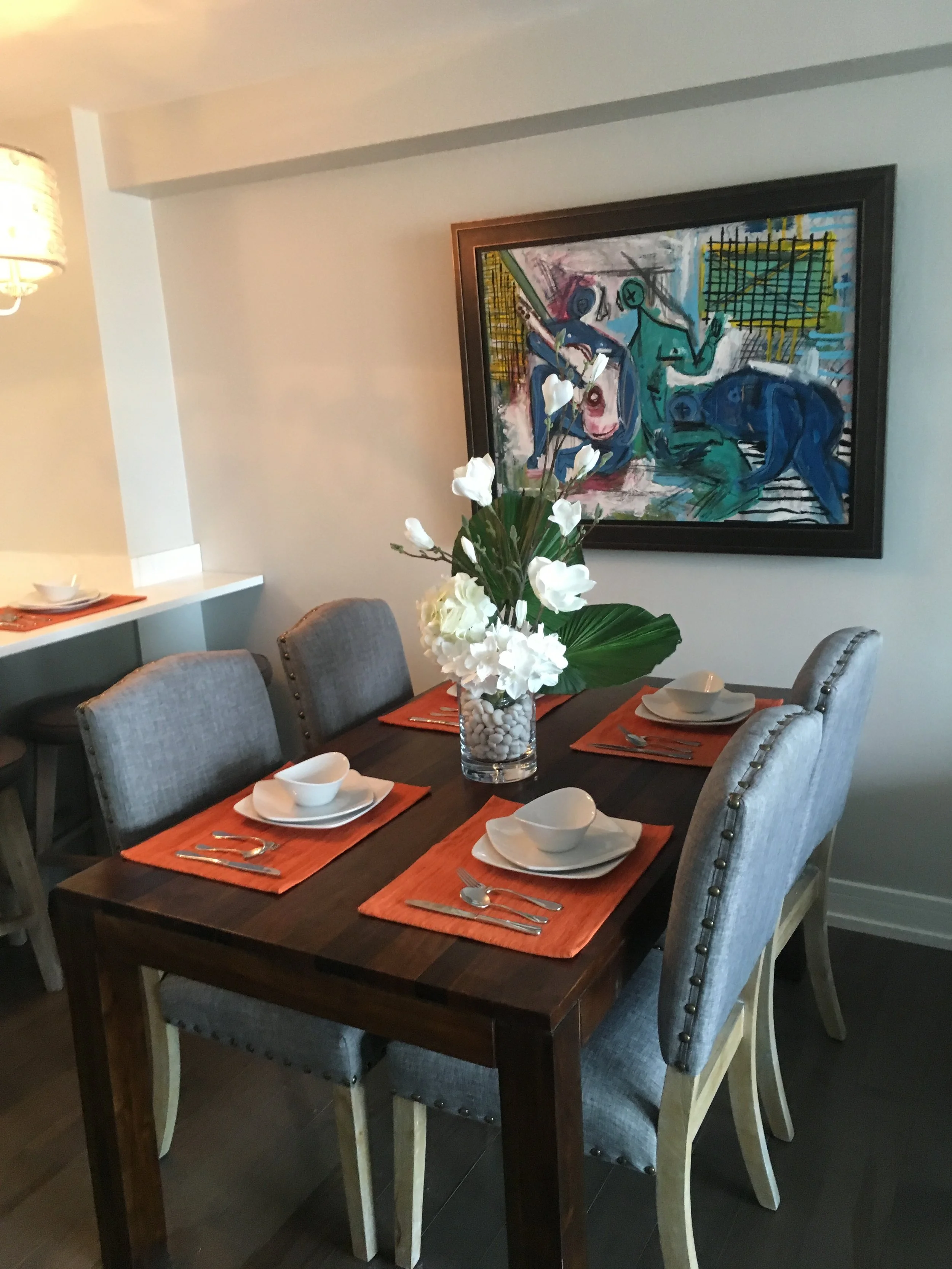 A dining room table set with white bowls and silverware on orange placemats, a vase with white flowers as a centerpiece, surrounded by gray upholstered chairs, with a colorful abstract painting on the wall in the background.