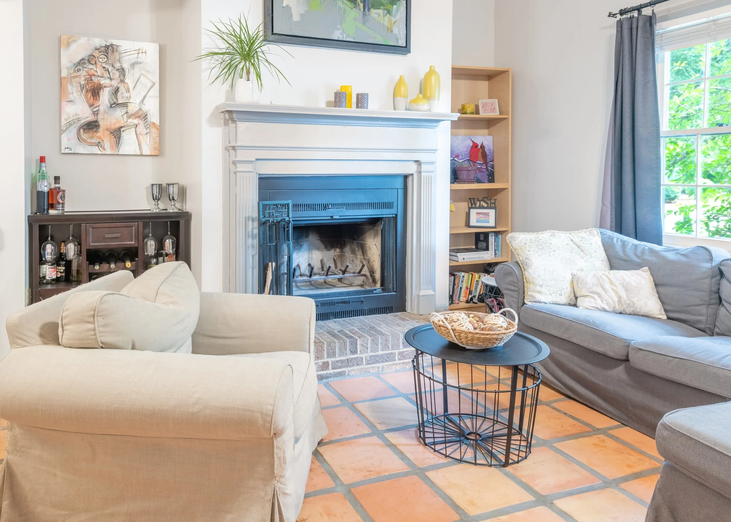 A cozy living room with a fireplace, a beige armchair, a gray sofa with decorative pillows, a black round coffee table with a wireframe base, a wooden bookcase with artwork and decorative items, and a window with blue curtains and lush green trees ou
