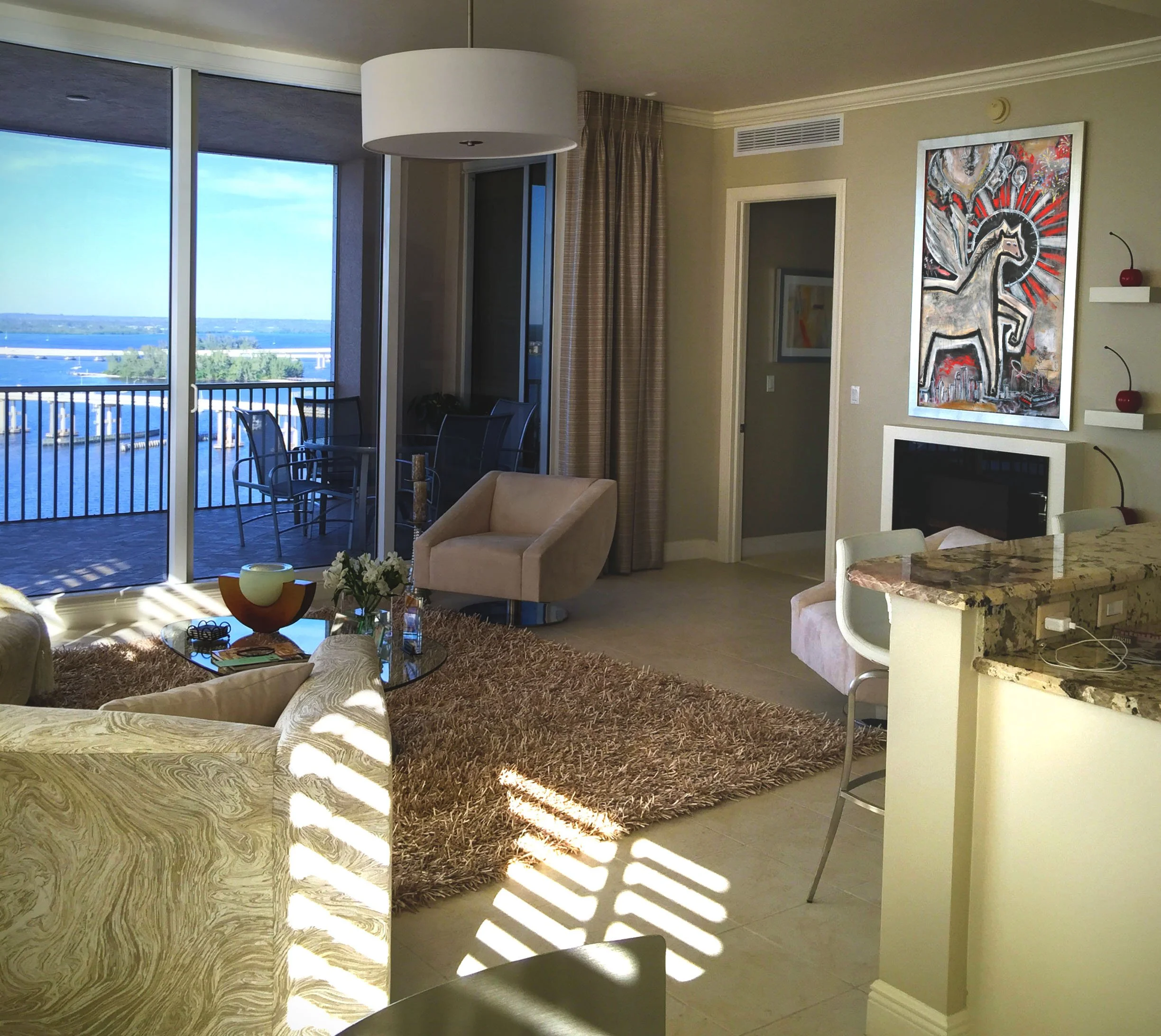 A living room with large windows showing a view of water, with balcony and outdoor chairs. Inside, there is a beige armchair, a glass coffee table with decorative items, a shaggy brown rug, a wall-mounted TV, a colorful abstract horse painting, and a