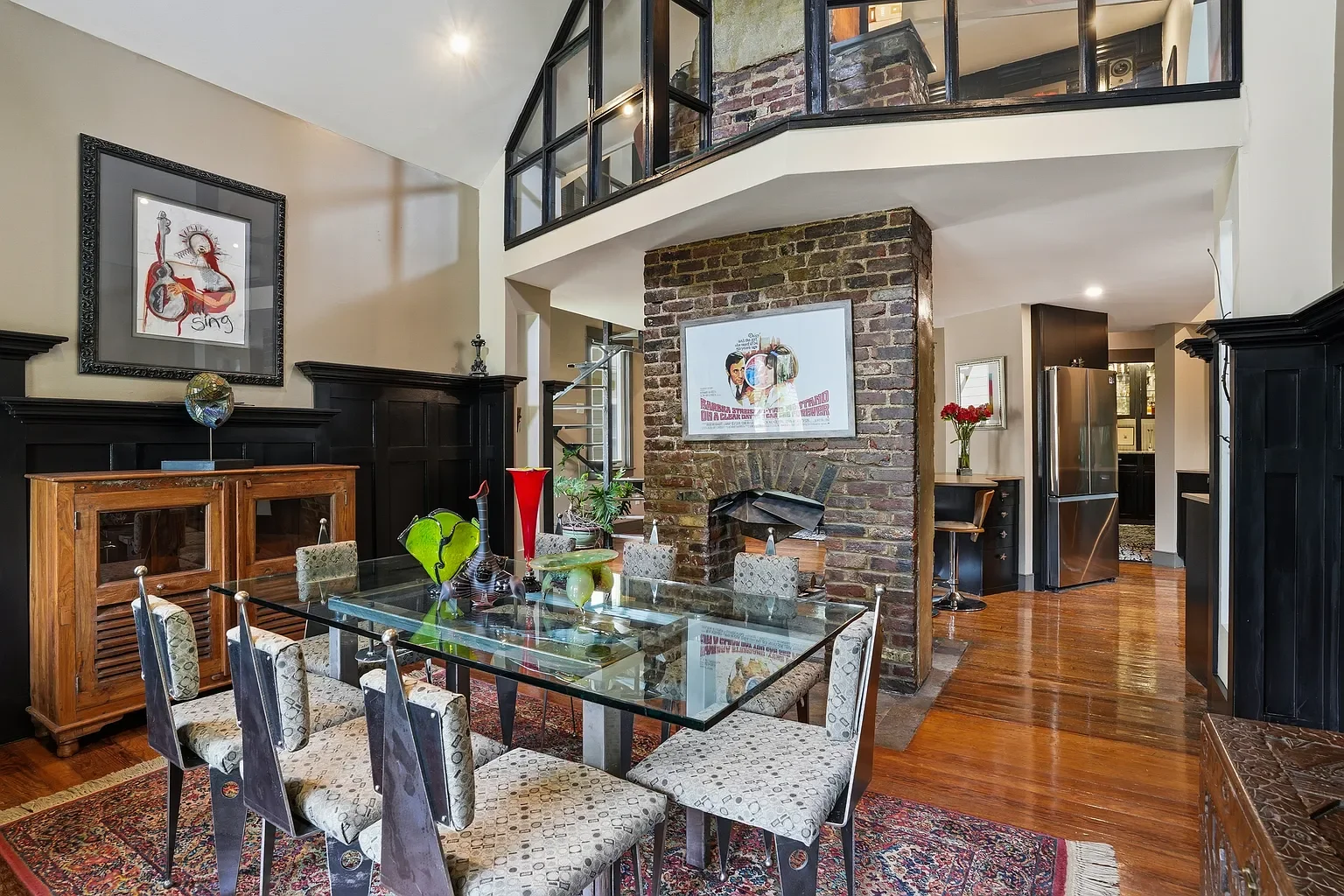 Interior view of a dining room with a glass table, patterned chairs, a brick fireplace, dark wood wall paneling, and artwork on the walls. There are decorative items and flowers on the table and shelves, with a staircase and a kitchen area visible in