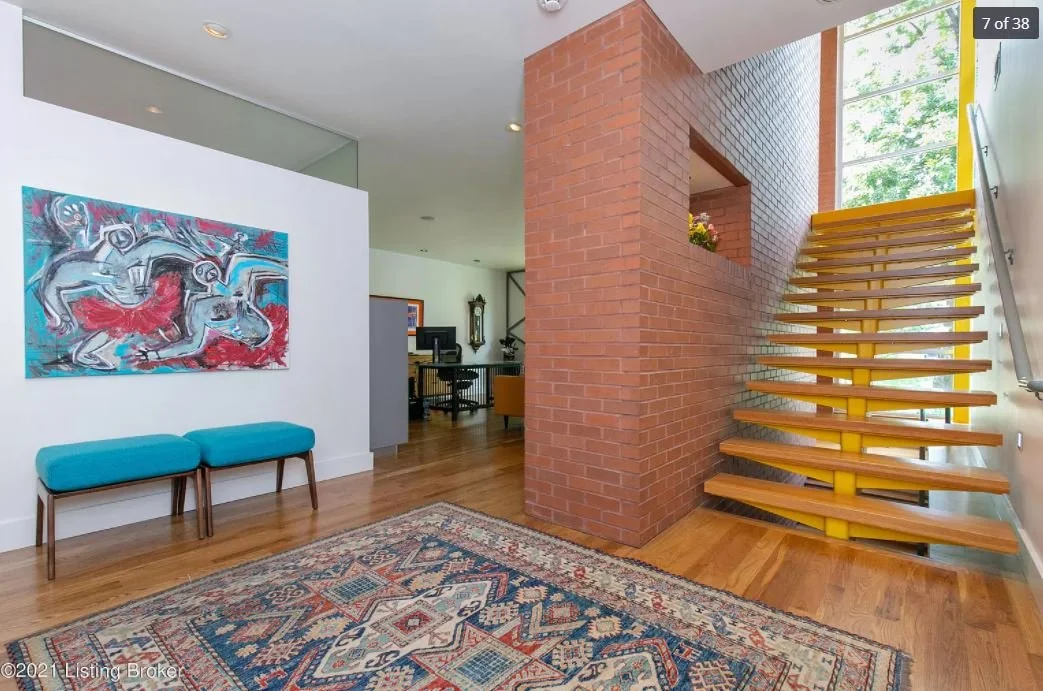 Modern interior of home with wooden staircase, brick wall, colorful abstract artwork, and a patterned area rug on hardwood floor.