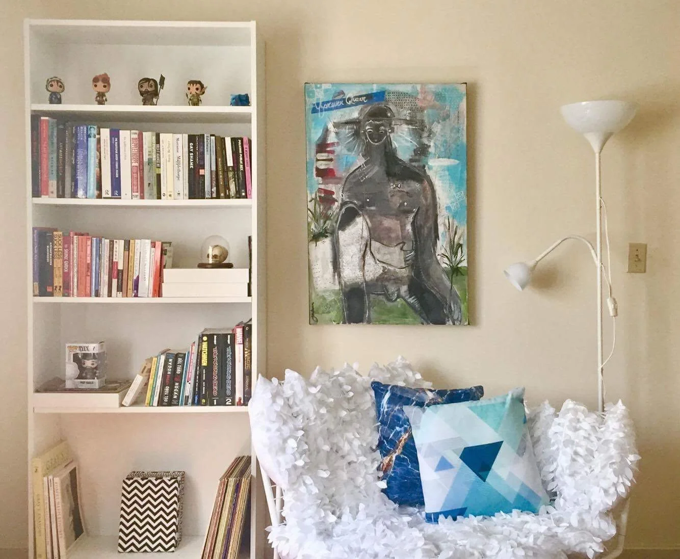A cozy corner of a room with a white bookshelf filled with books and decorative items, a colorful abstract painting on the wall, a floor lamp, and a small white chair with a blue geometric pillow and white decorative paper flowers.
