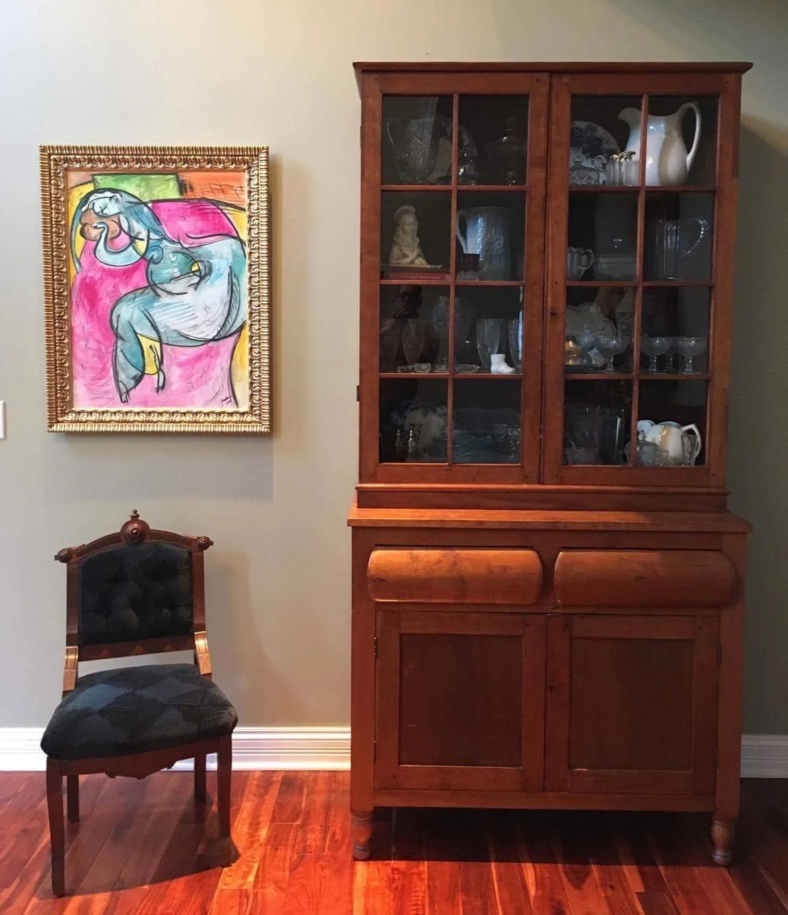 A living room corner with a colorful abstract painting of a horse on the wall, a wooden display cabinet with glass doors filled with glassware and decorative items, and a dark upholstered chair with a wooden frame on a polished wood floor.