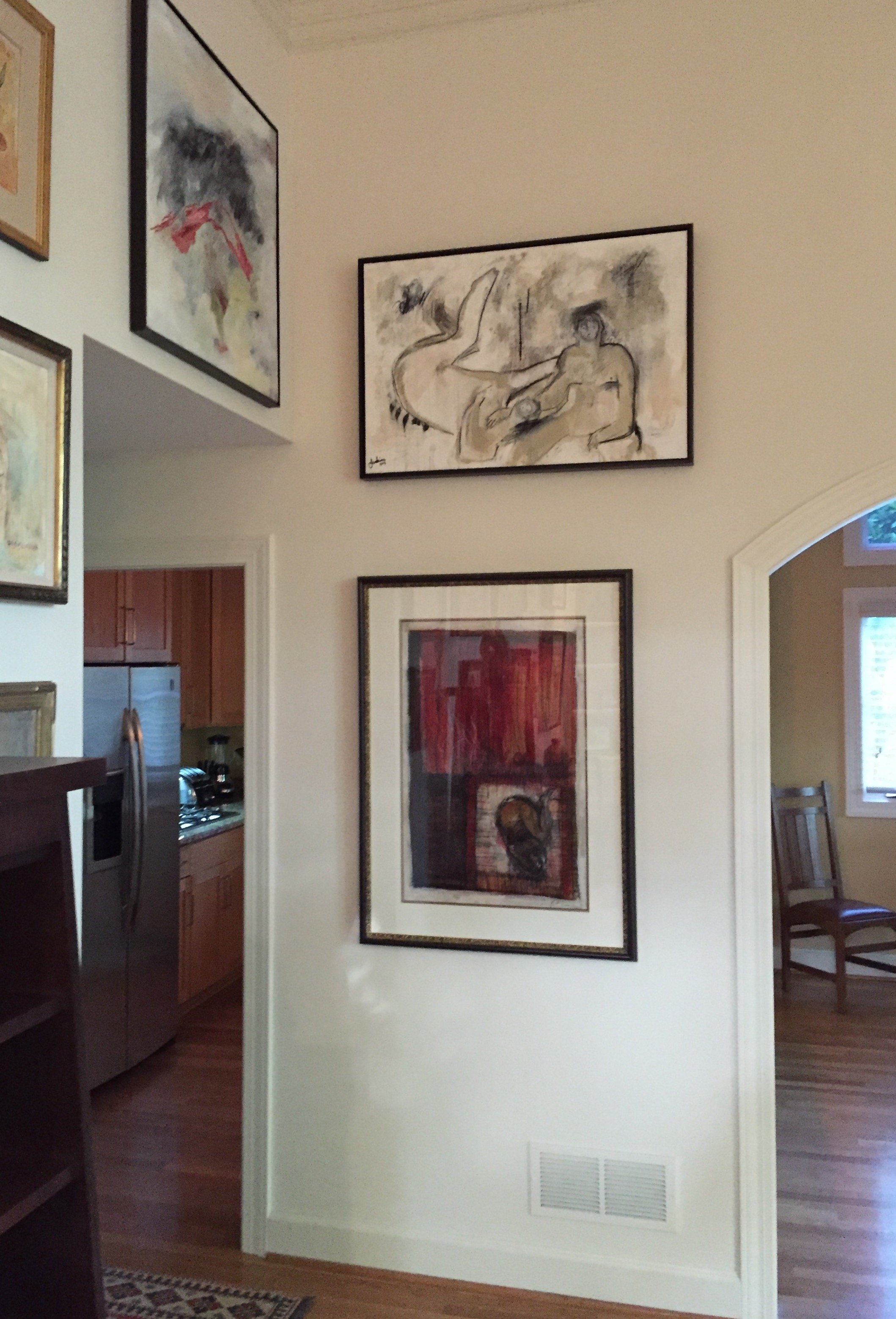 Living room with framed artwork on cream-colored wall, open kitchen with wooden cabinets and stainless steel appliances, hardwood floors, and a wooden chair near a window.