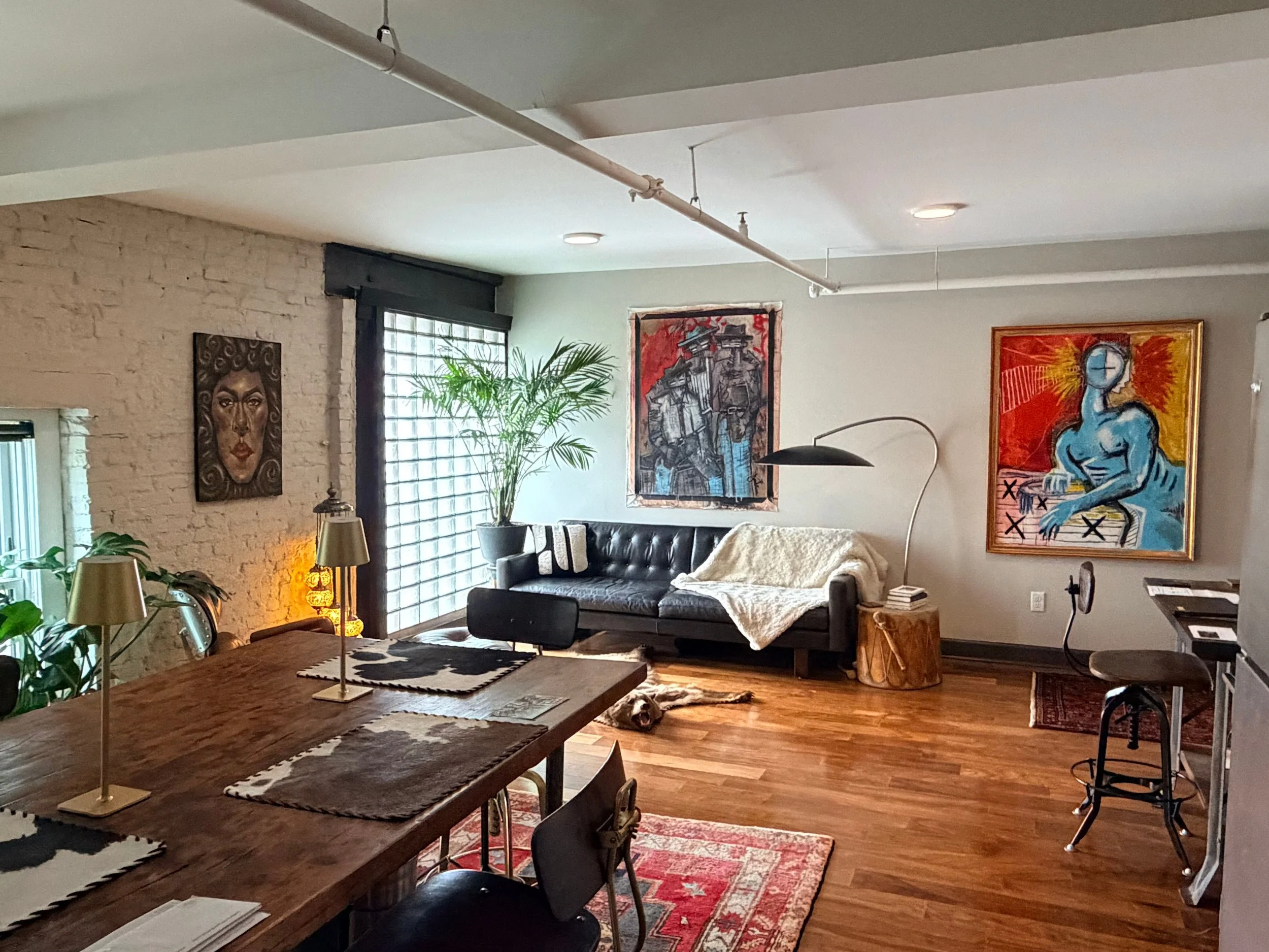 A cozy living room with exposed brick walls, colorful artwork, a black leather sofa, and a large wooden dining table with decorative lamps, plants, and patterned rugs. Paintings featured are by Louisville, KY artist Joshua Jenkins.
