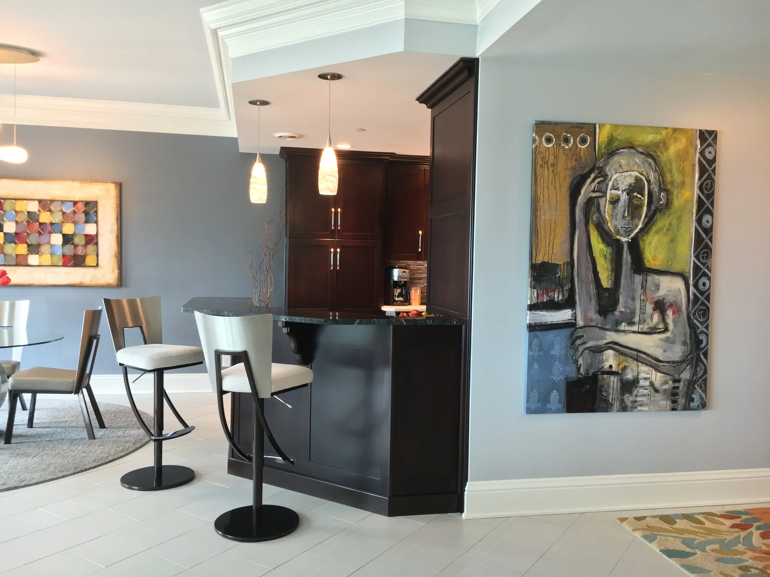 A modern kitchen and dining area with a dark wood kitchen cabinet, a granite countertop, and artistic paintings on the wall. There are pendant lights hanging from the ceiling and white chairs around a glass table.