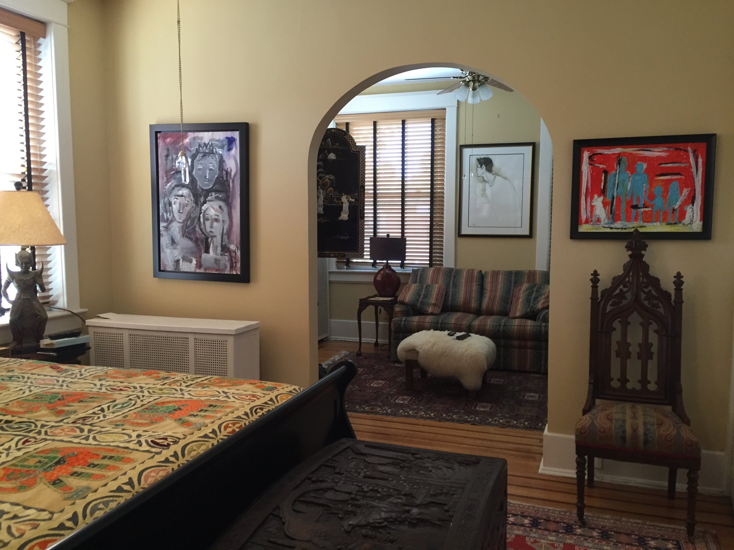 Living room with yellow walls, framed artwork, wooden blinds, a ceiling fan, a couch with striped upholstery, a small table with a red vase, and a decorative wooden chair.