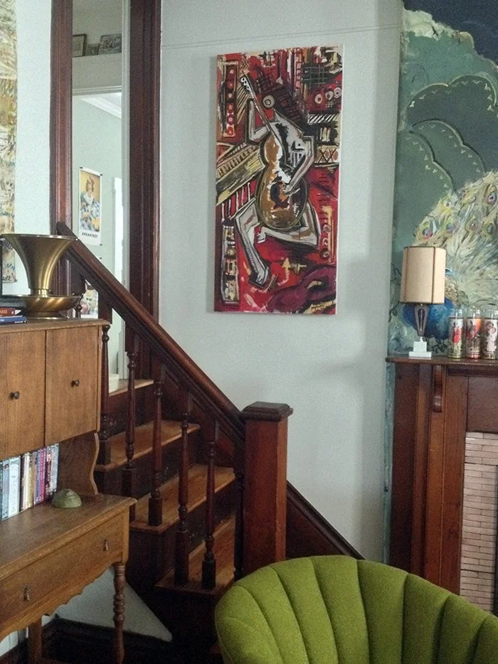 Interior of a home with a wooden staircase, mid-century furniture, a green upholstered chair, and colorful artwork on the wall, including a painting of a guitar.
