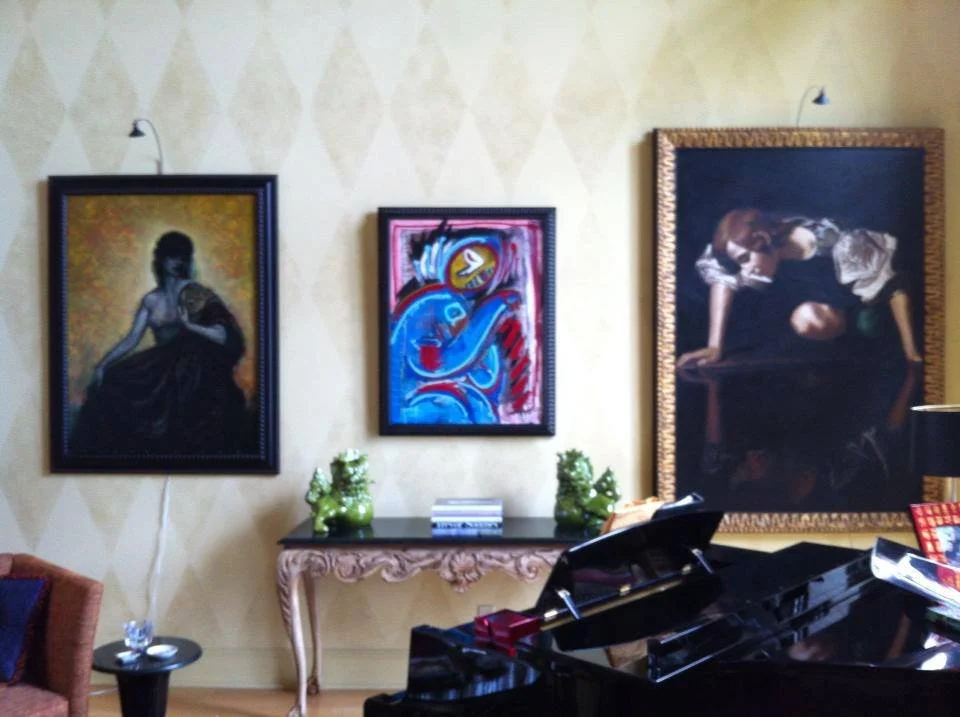 A living room with three framed paintings on a beige patterned wall, a black grand piano, and a decorative console table with green vases and books.
