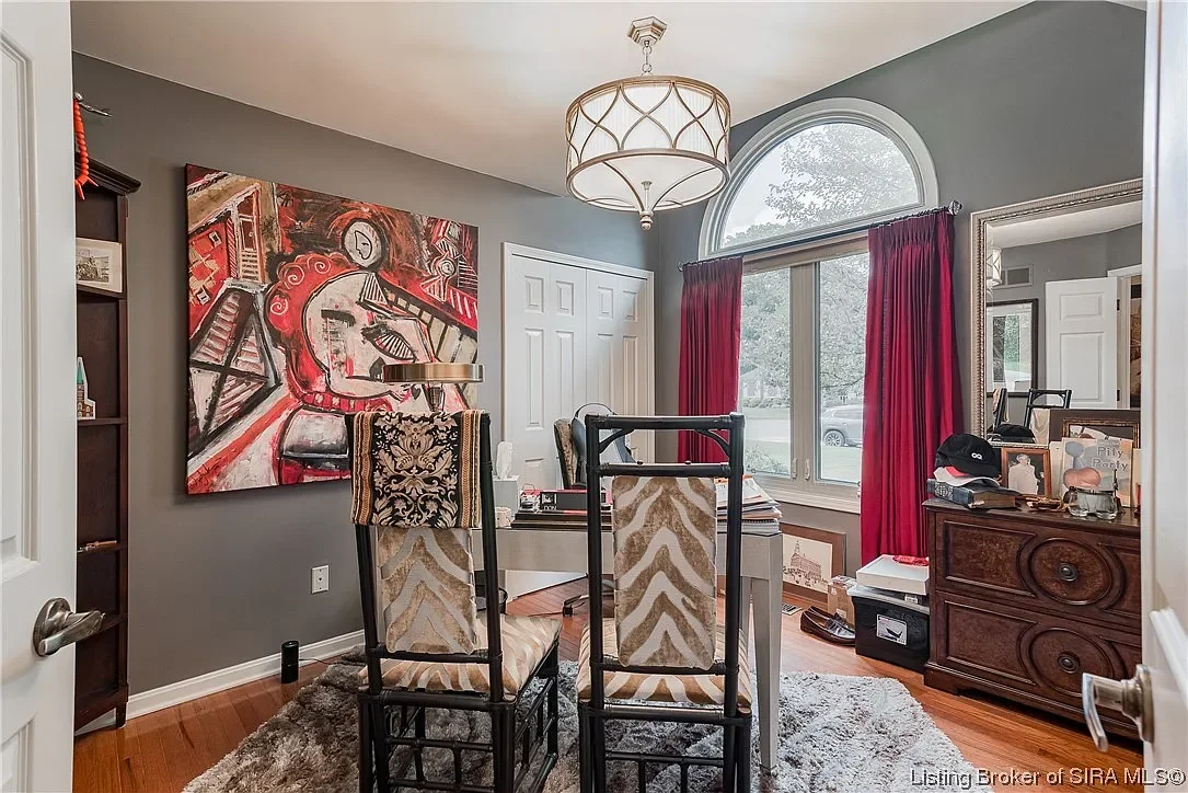 A home office with gray walls, a large painting of a woman playing the guitar, a white desk with chairs, a window with red curtains, a dresser, and various decorative items.