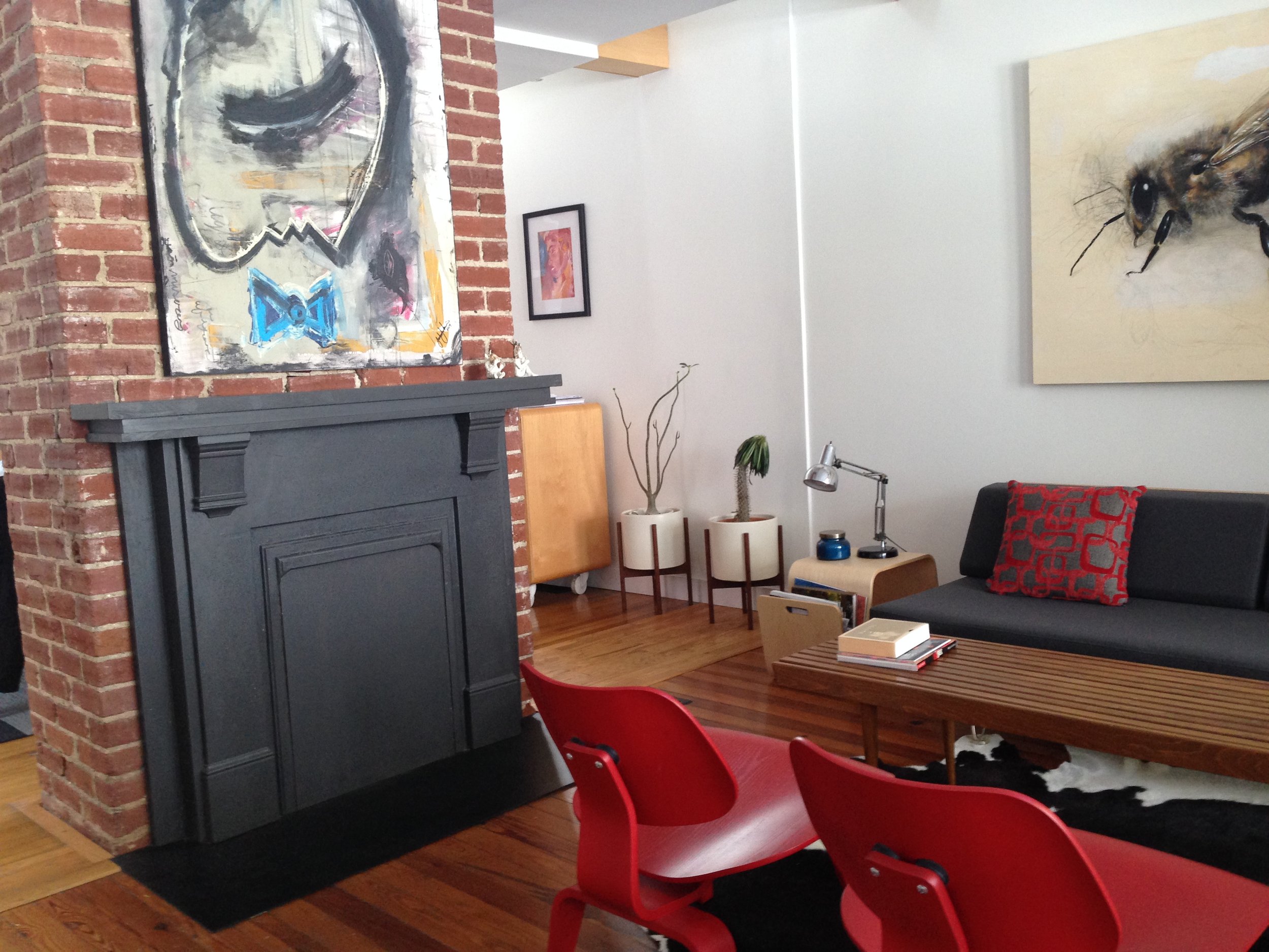 Living room with exposed brick wall, artwork, black fireplace, two potted plants, red chairs, wooden table, gray couch, and wall-mounted artwork.