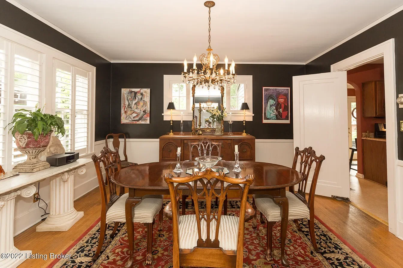 A formal dining room with a wooden oval table and six matching chairs. A chandelier hangs above. The room has dark walls with white trim, windows with shutters, and decorative framed artwork. A sideboard with a mirror, lamps, and floral decor is at t