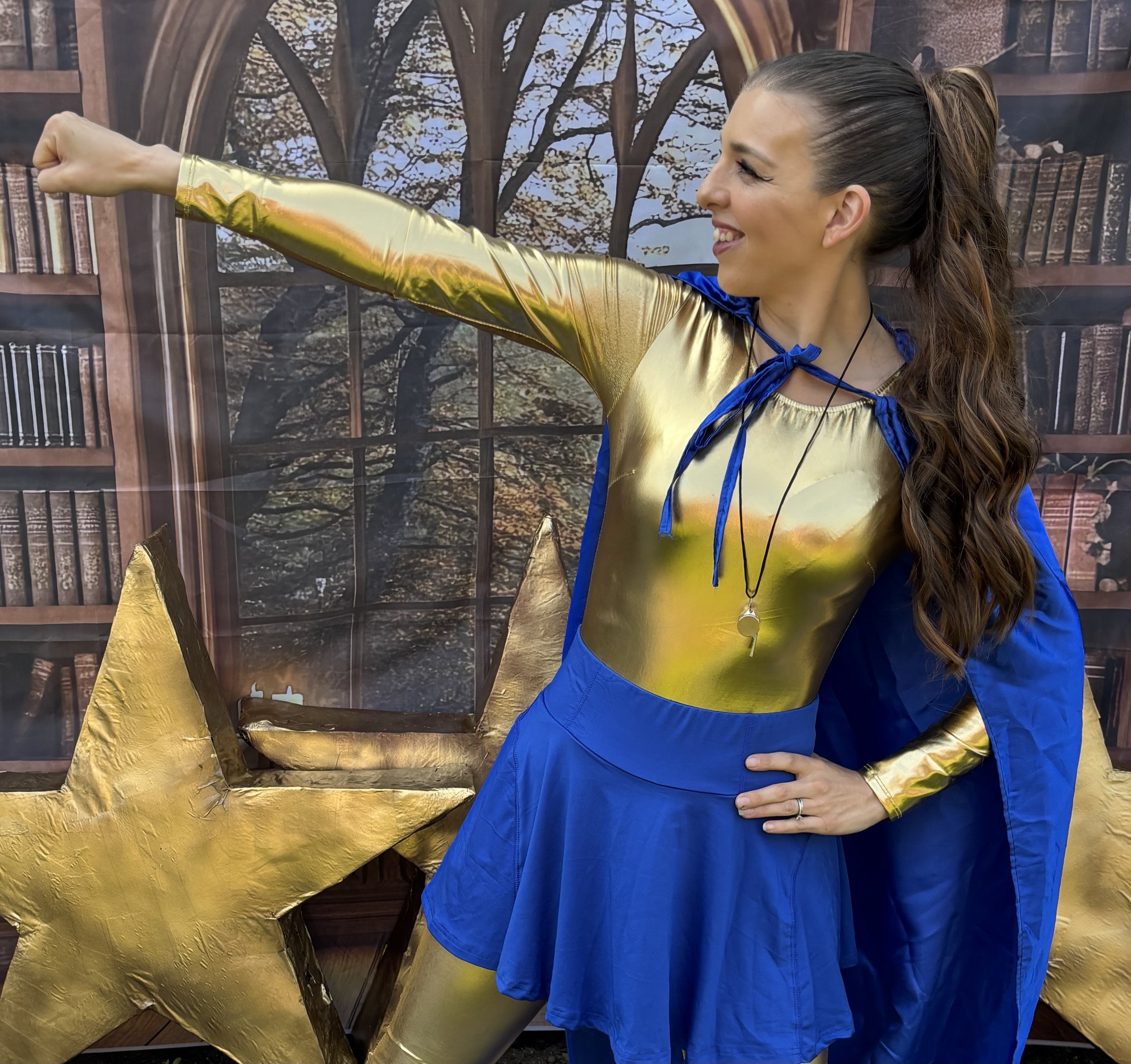 Woman dressed as a superhero in a shiny gold and blue costume, smiling and gesturing heroically with a large gold star prop in the background.