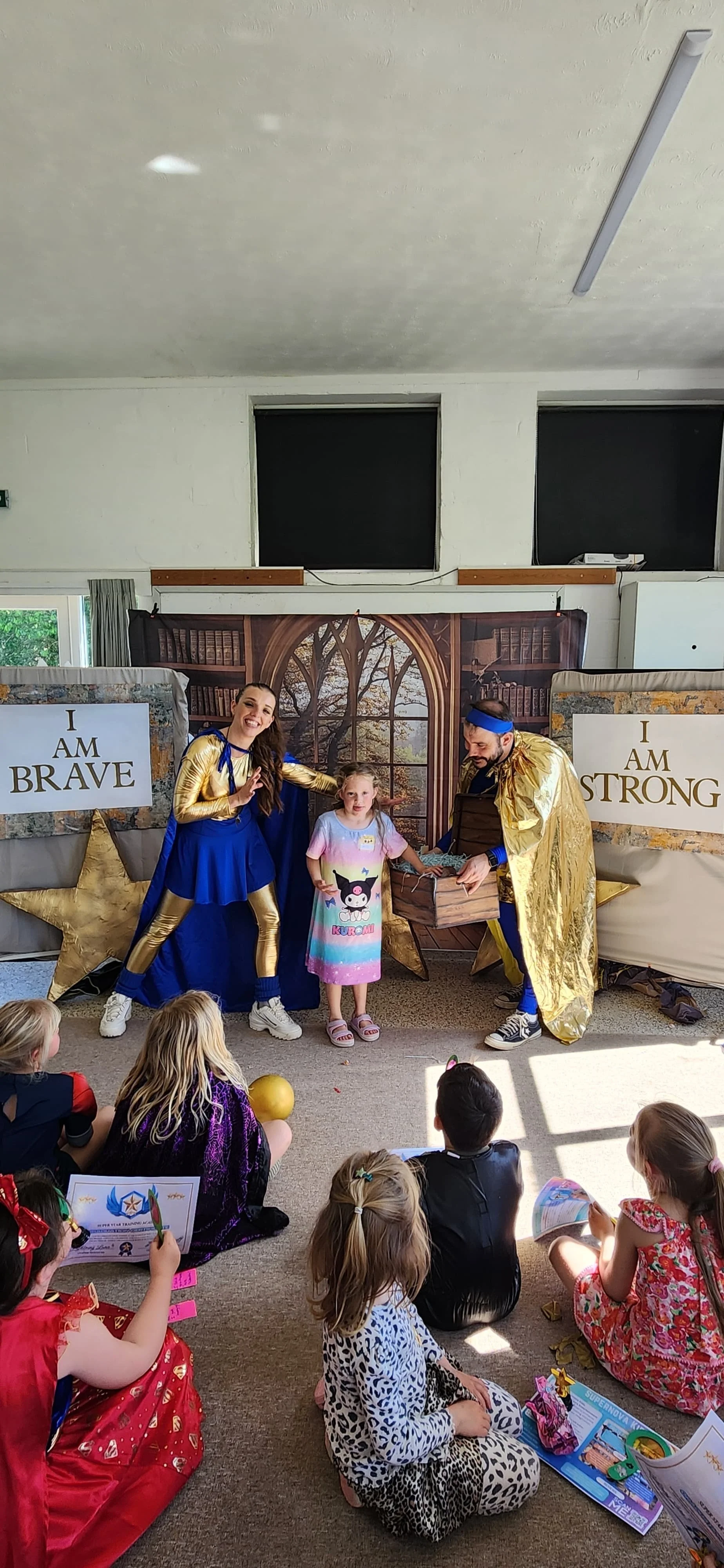 Children watching a magic show with two performers dressed as superheroes, one female and one male, in a room decorated with signs saying 'I AM BRAVE' and 'I AM STRONG,' and a backdrop featuring a window and trees.