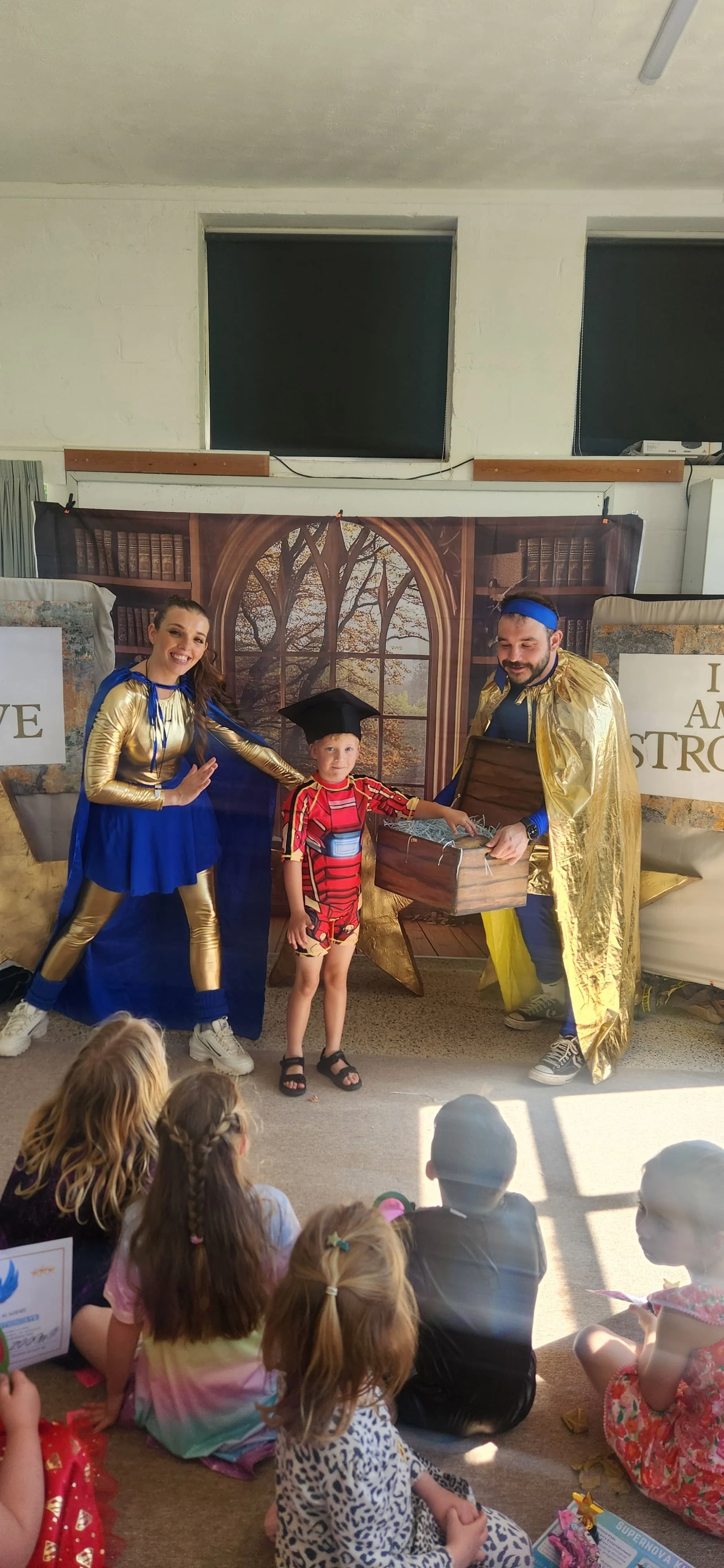 Children watching a puppet show with two performers dressed as superheroes, one in a gold and blue costume and the other in a gold and blue cape, holding a treasure chest.
