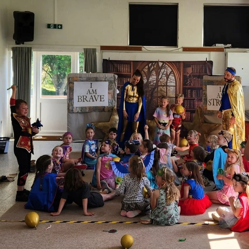 Children dressed in colorful costumes participating in a classroom play or event with characters portraying bravery and strength, featuring signs that read 'I AM BRAVE' and 'I AM STRONG'.