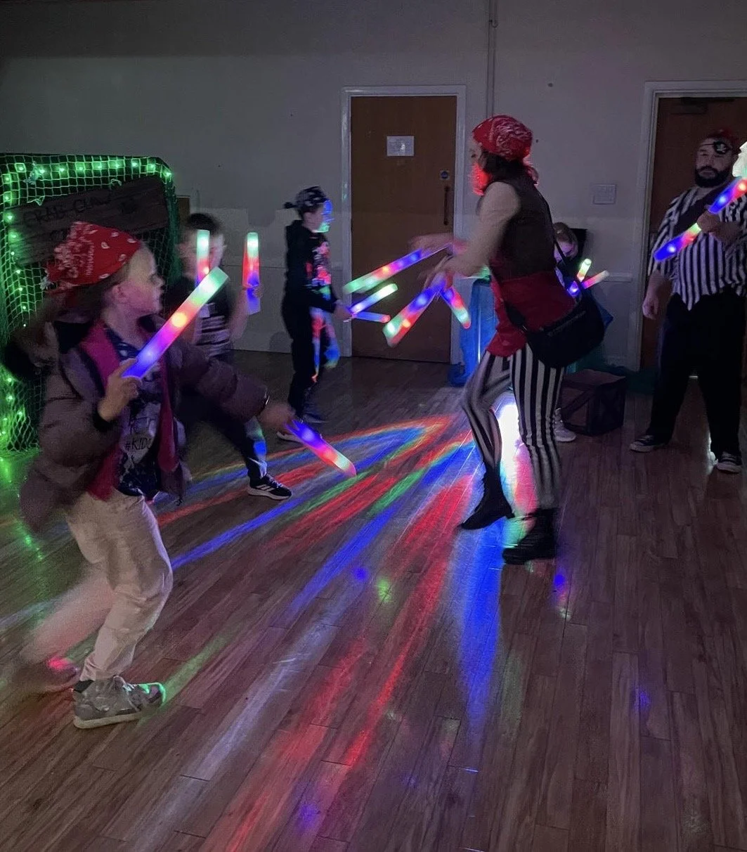 Children dancing with LED glow sticks at a party, under a green-lit hockey goal net in a dimly lit room.
