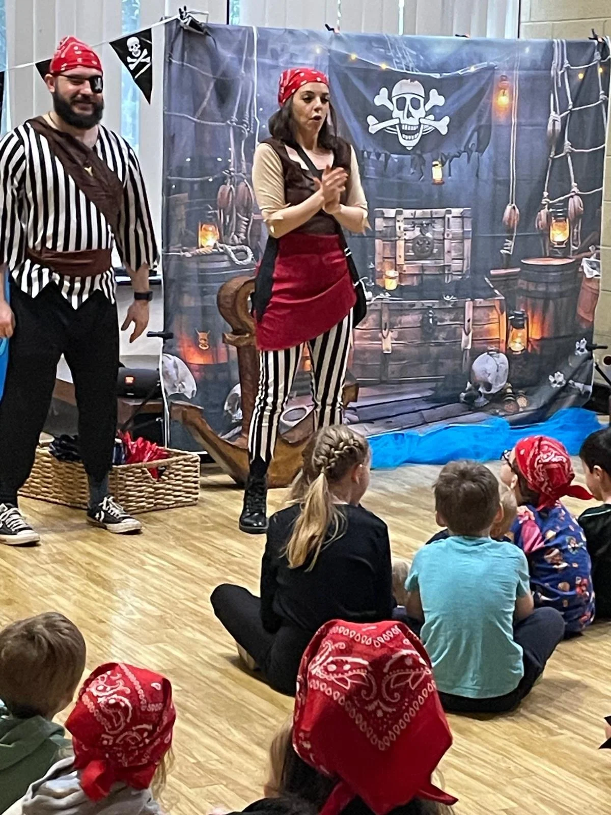 Pirate-themed theatrical performance for children, featuring a woman and man in pirate costumes with a painted backdrop of a pirate ship's interior, children sitting on the floor during a pirate party watching, some wearing red bandanas.