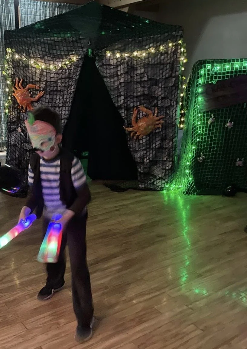 A child wearing a colourful mask and a striped shirt holding glow sticks in front of a Halloween-themed backdrop with large plastic crabs and decorative netting. This is a scene from one of our recent birthday parties in Bournemouth.