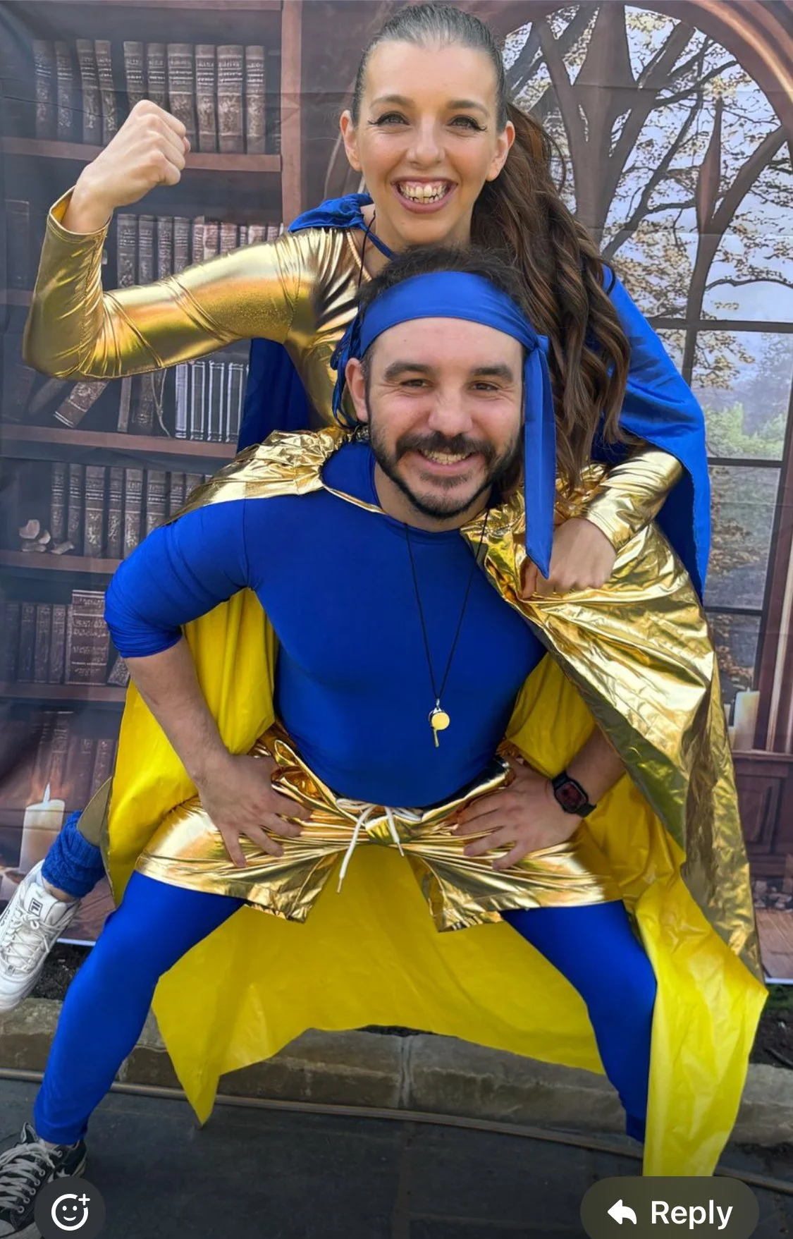 Two adults dressed in superhero costumes, with blue and gold outfits, pose in front of a backdrop resembling a library. The woman is sitting on the man's shoulders, flexing her arm and smiling, while the man stands with hands on his hips, also smiling.