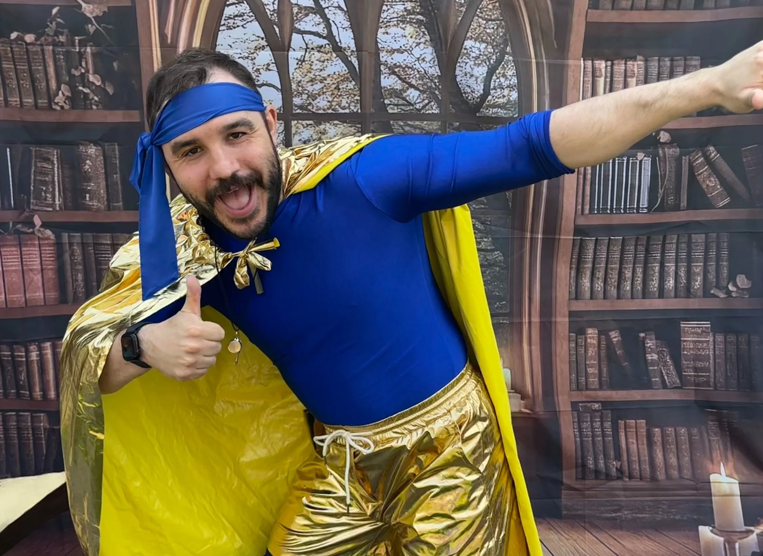 A man dressed as a superhero in a blue and gold costume posing enthusiastically in front of a bookshelf backdrop, giving a thumbs-up.