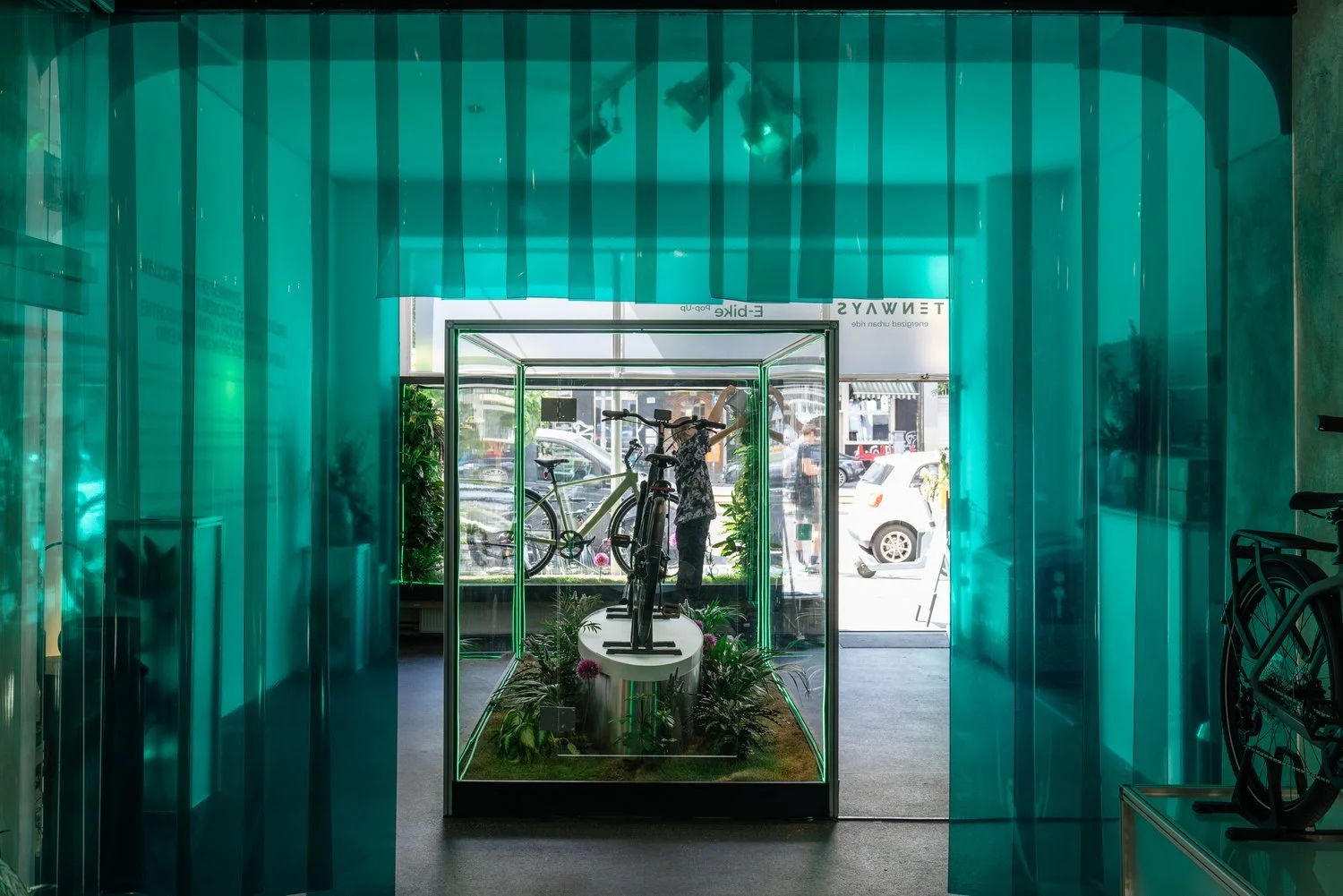 Pop Up shop in Berlin by Culture Defined for Tenways Inside a bicycle shop with a display case featuring bicycles, surrounded by teal-colored walls and curtains, with large windows showing cars outside.