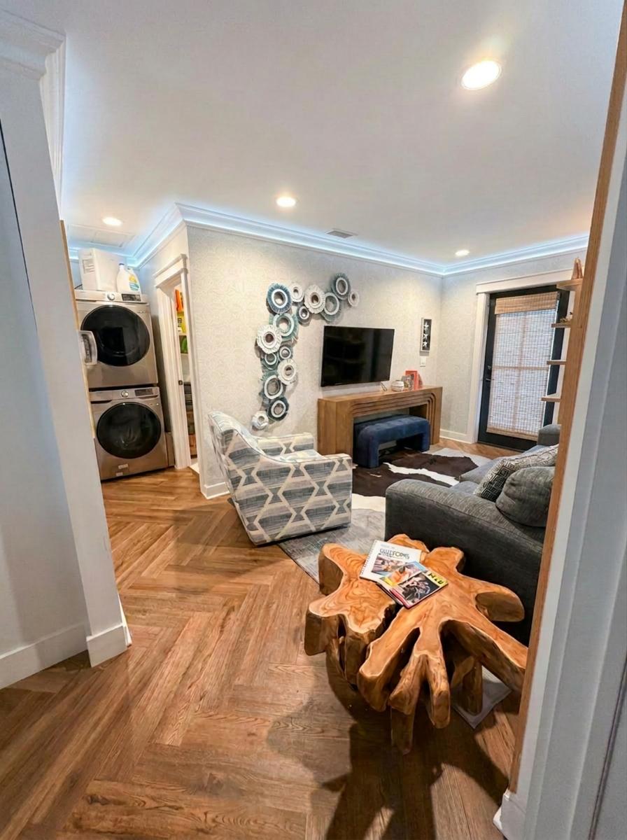 Living room with patterned armchair, gray sofa, wooden coffee table, wall TV, decorative wall art, and laundry area with stacked washer and dryer.