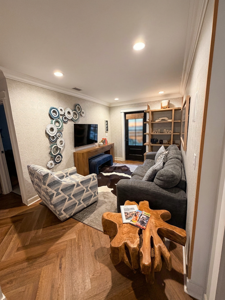 A cozy living room with a patterned armchair, a grey sofa with cushions, a wooden side table with magazines, a cowhide rug, a TV on a wooden stand, decorative wall art, and a bookshelf, with hardwood floors and ceiling lights.