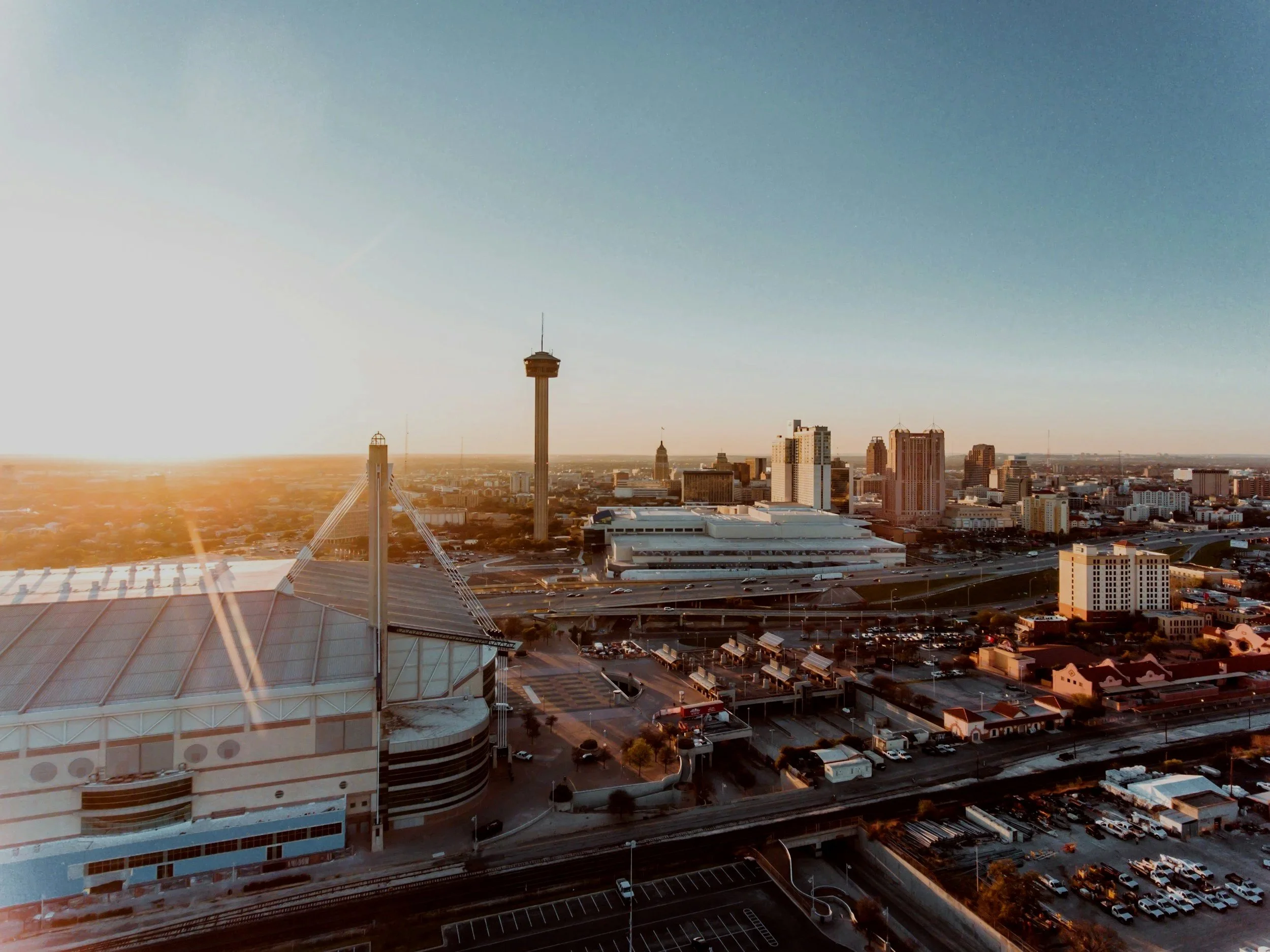 A city skyline at sunset with tall buildings, a prominent tower, and a shopping mall in the foreground.