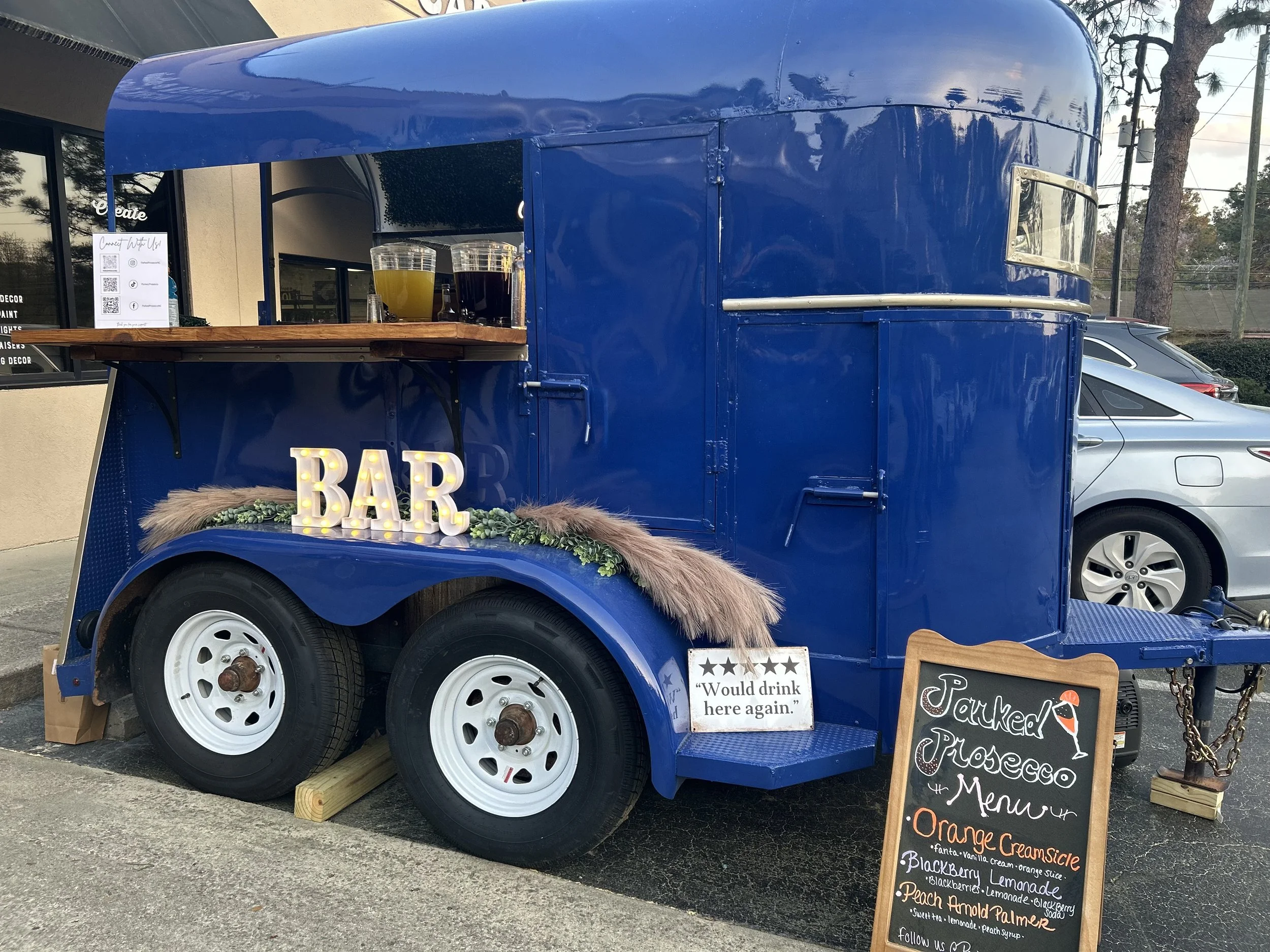 Outdoor event with a blue mobile bar trailer named 'Parked Prosecco.' The setup includes string lights, bartenders, and guests seated at tables. Tents in the background with people dining and socializing on a lawn.