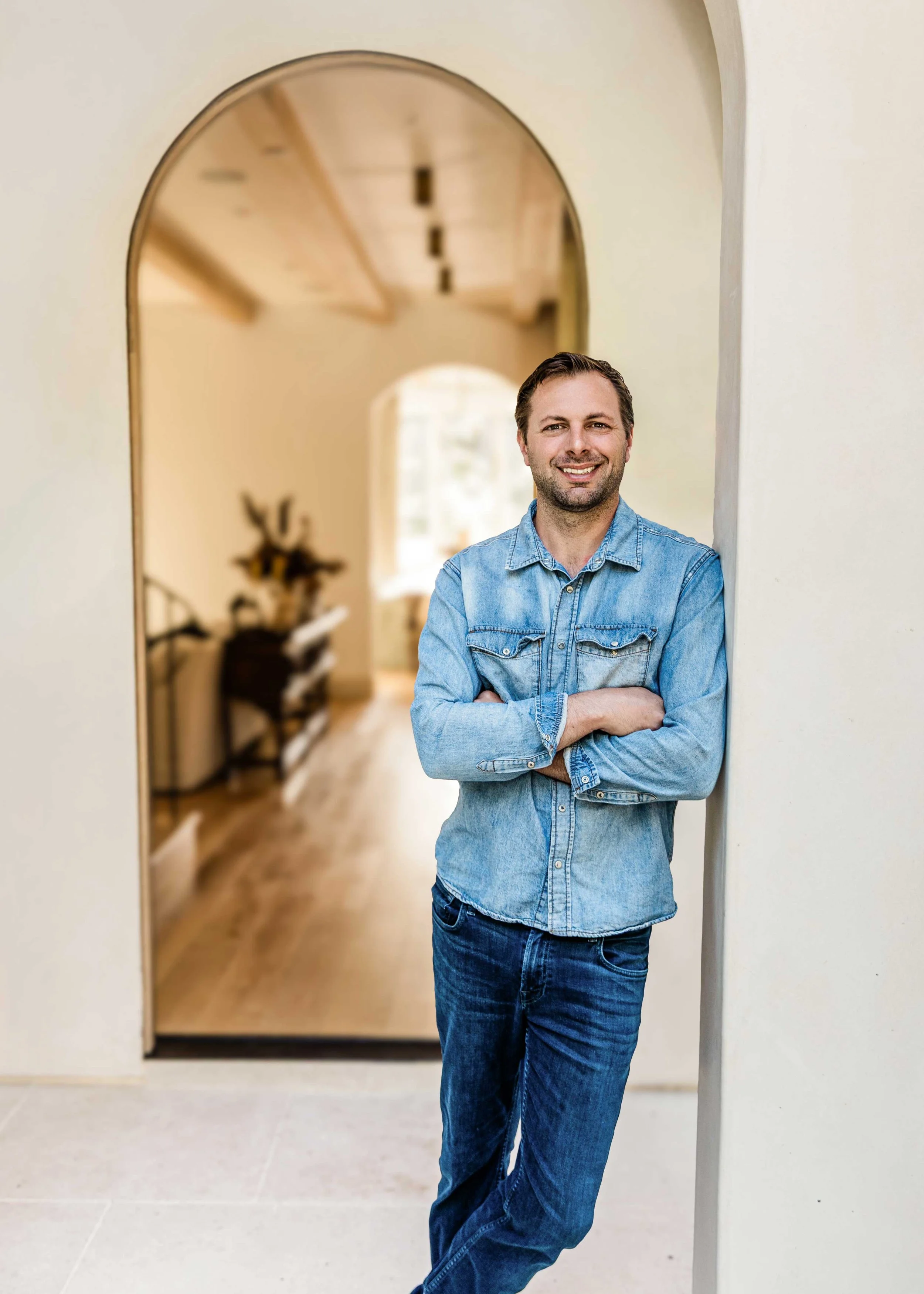 A man with short brown hair and a beard, wearing a denim shirt and jeans, standing with arms crossed and smiling in a home interior with an arched doorway and wooden flooring.