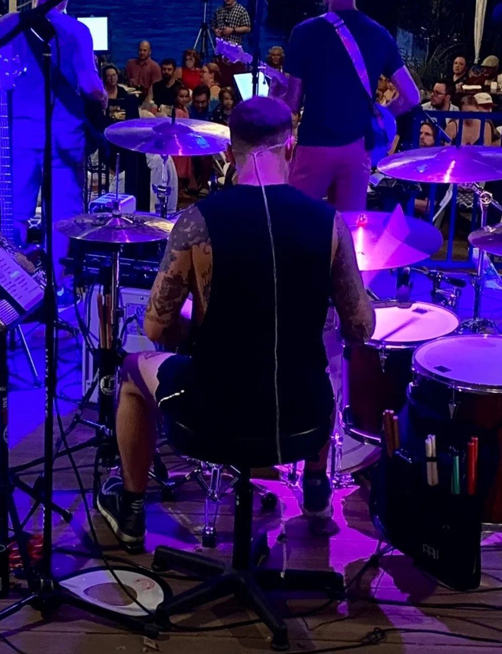 Musicians performing on stage with drums and guitar, audience watching in the background, purple stage lighting.