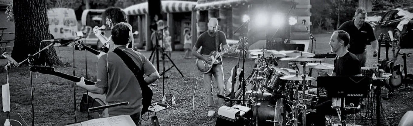 A group of musicians performing outdoors at night, with various instruments including guitars, drums, and microphones.