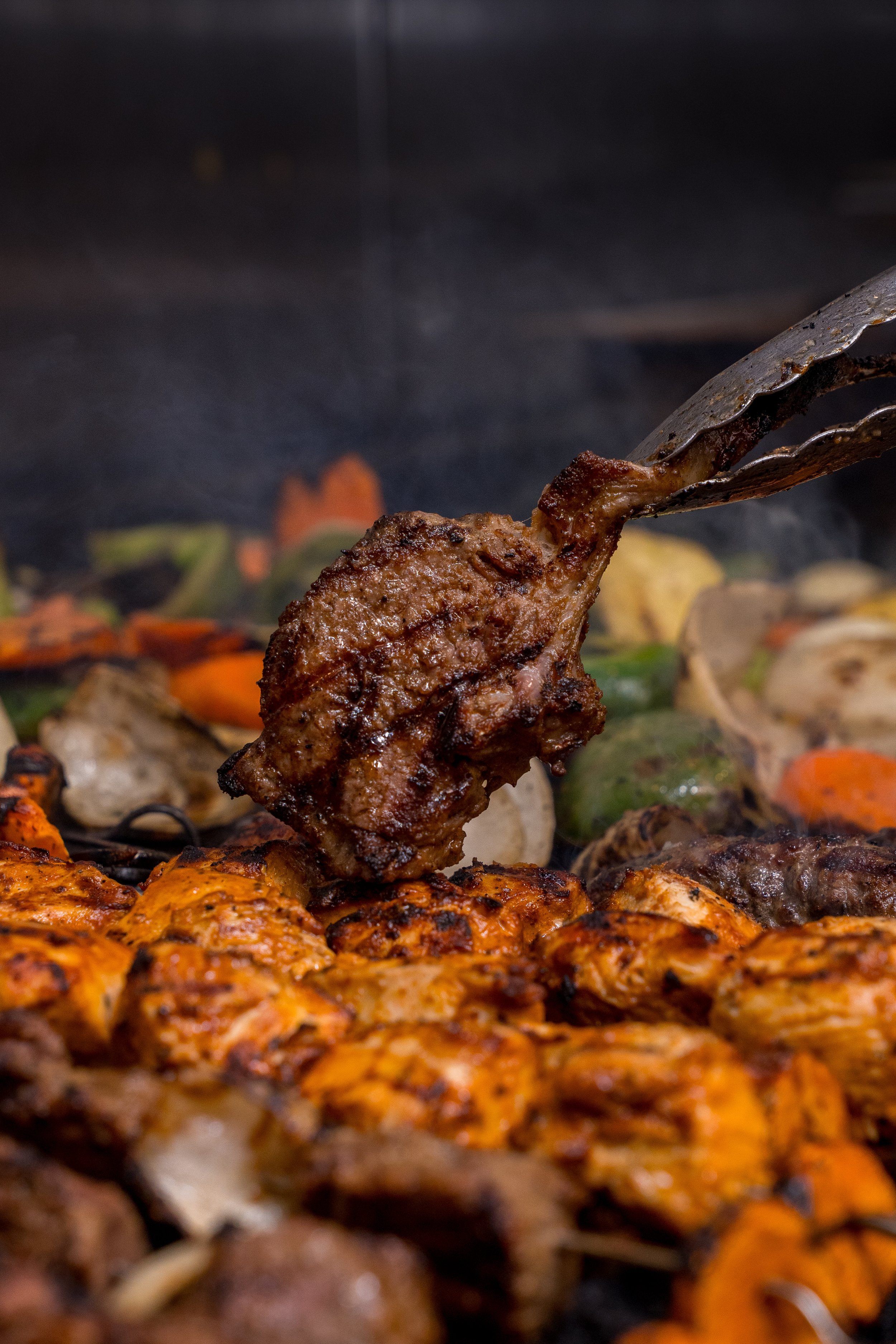 Close-up of grilled meat and vegetables on a barbecue with tongs holding a cooked piece of meat.