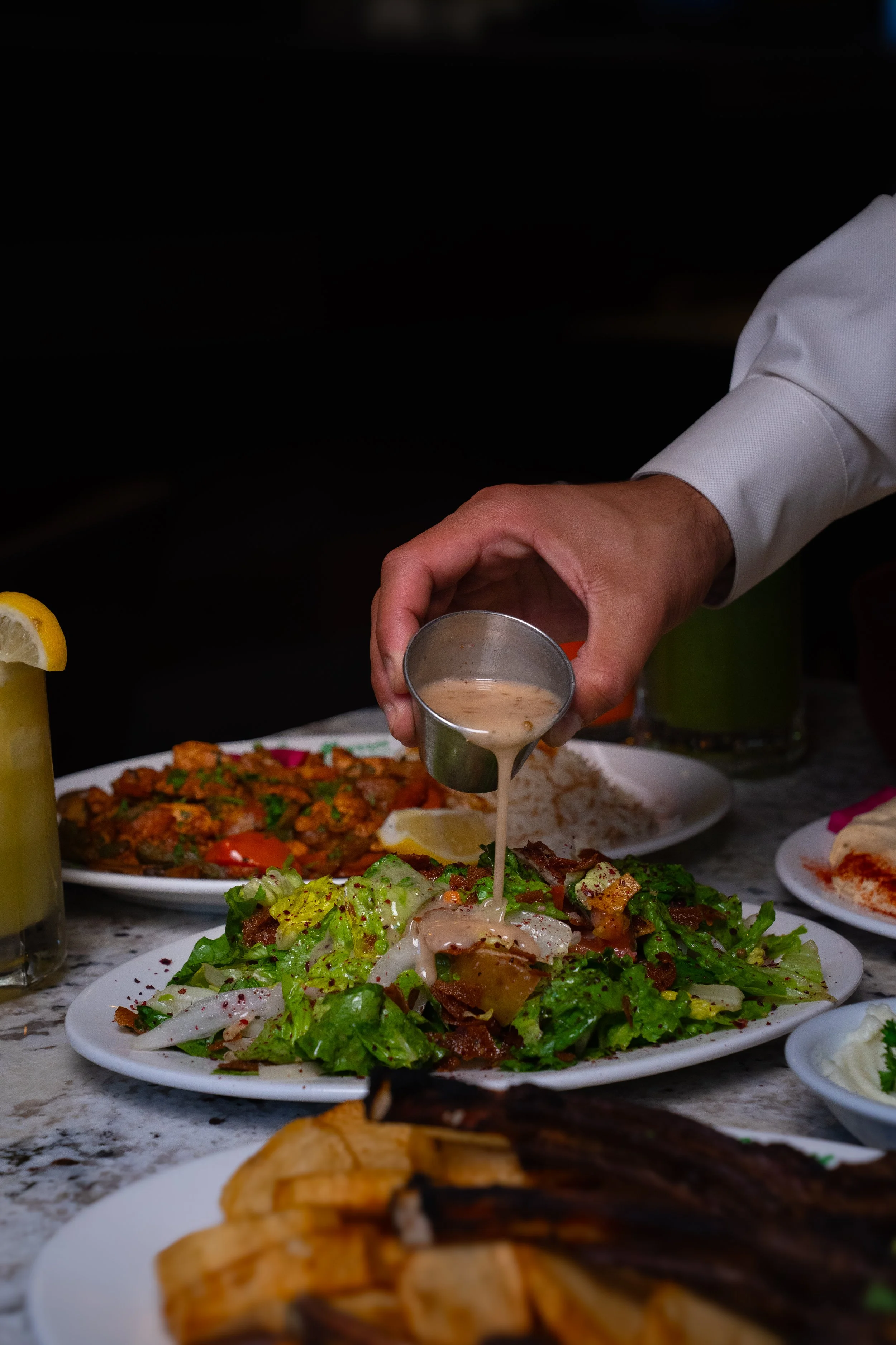 Person pouring dressing on salad, surrounded by plates of food and drinks on a table.