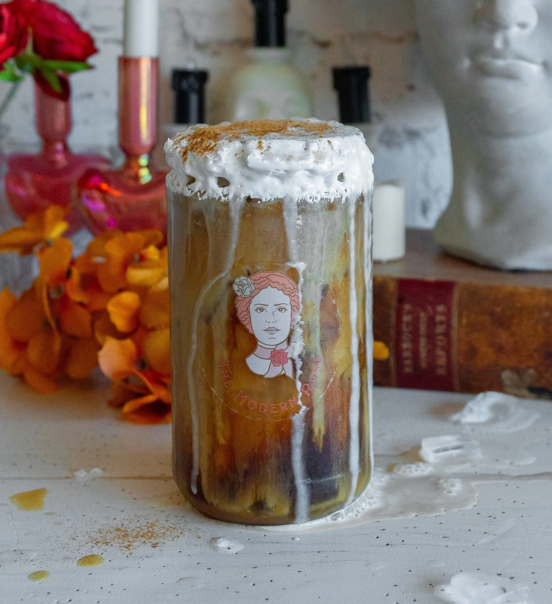 Iced coffee in a glass with whipped cream overflowing, decorated with autumnal elements and a vintage portrait, set on a rustic white table.