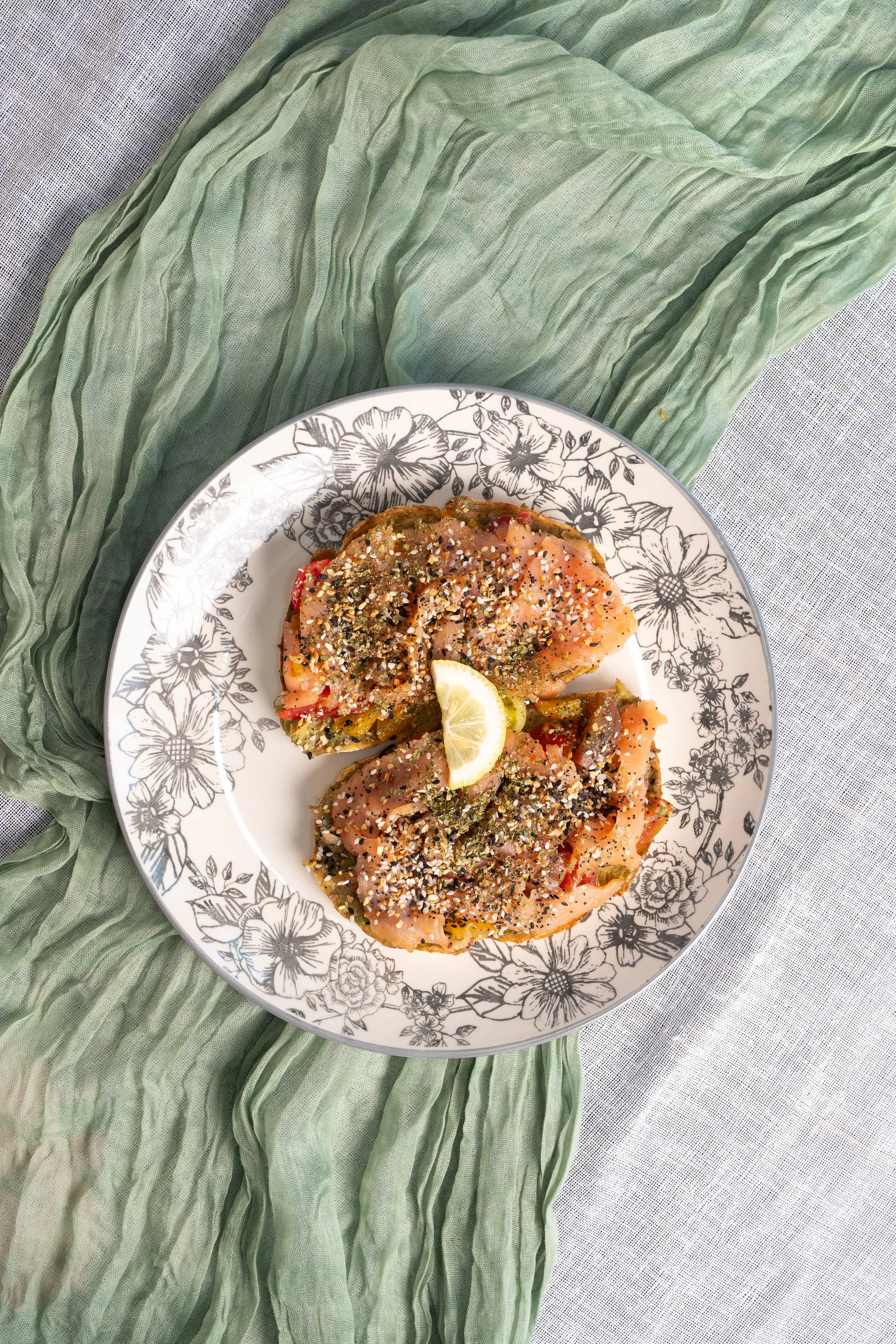 Plate with open-faced sandwiches topped with smoked salmon, seeds, herbs, and lemon wedge, on floral patterned dish and green cloth.