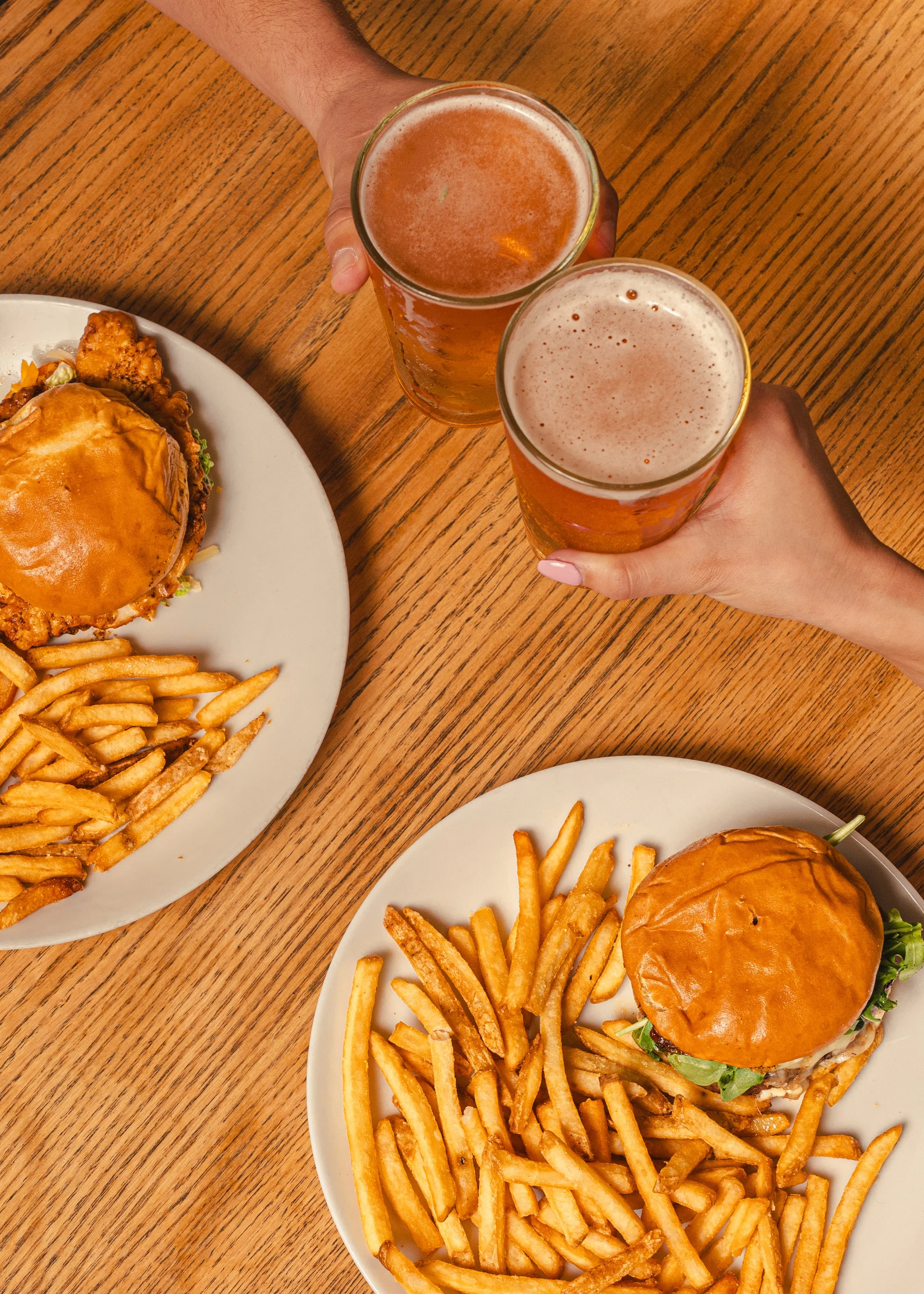 Two plates with burgers and fries on a wooden table, and two hands holding glasses of beer.